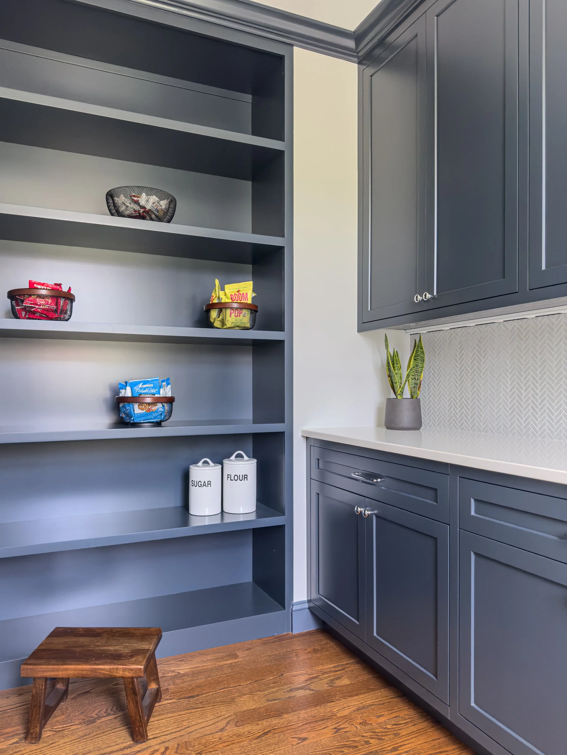Navy butlers pantry with floor to ceiling open shelving and closed cabinetry with a white quartz countertop and herringbone tile backsplash.