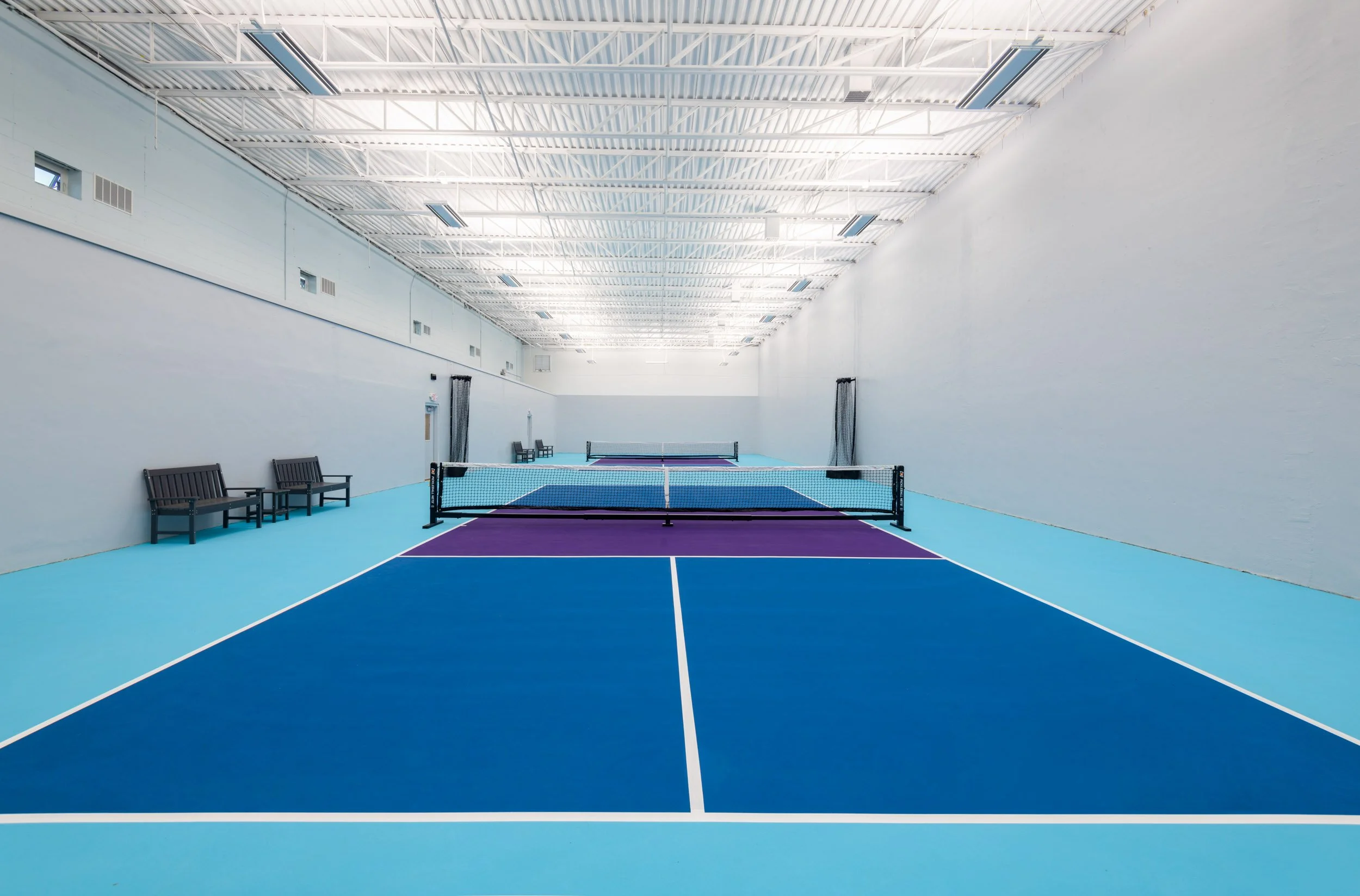 Indoor blue and purple pickleball courts with exposed steel i-joists and viewing area