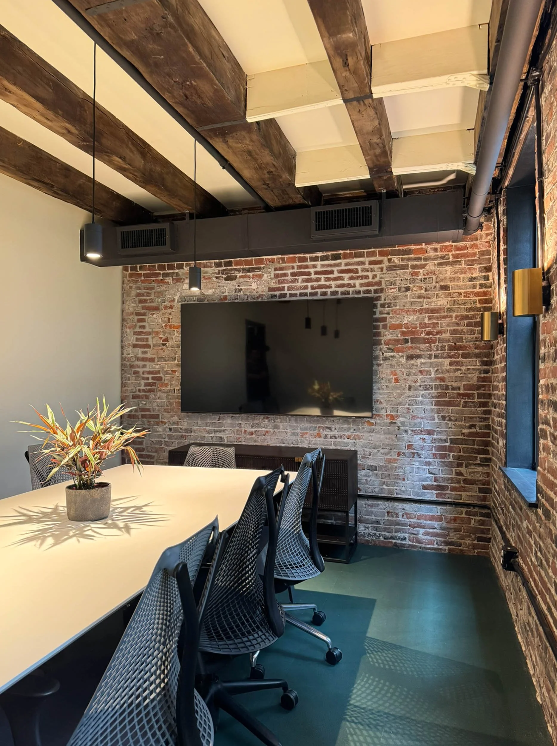 A small conference room with a wall-mounted television against exposed brick and original wood beams in a converted commercial space.