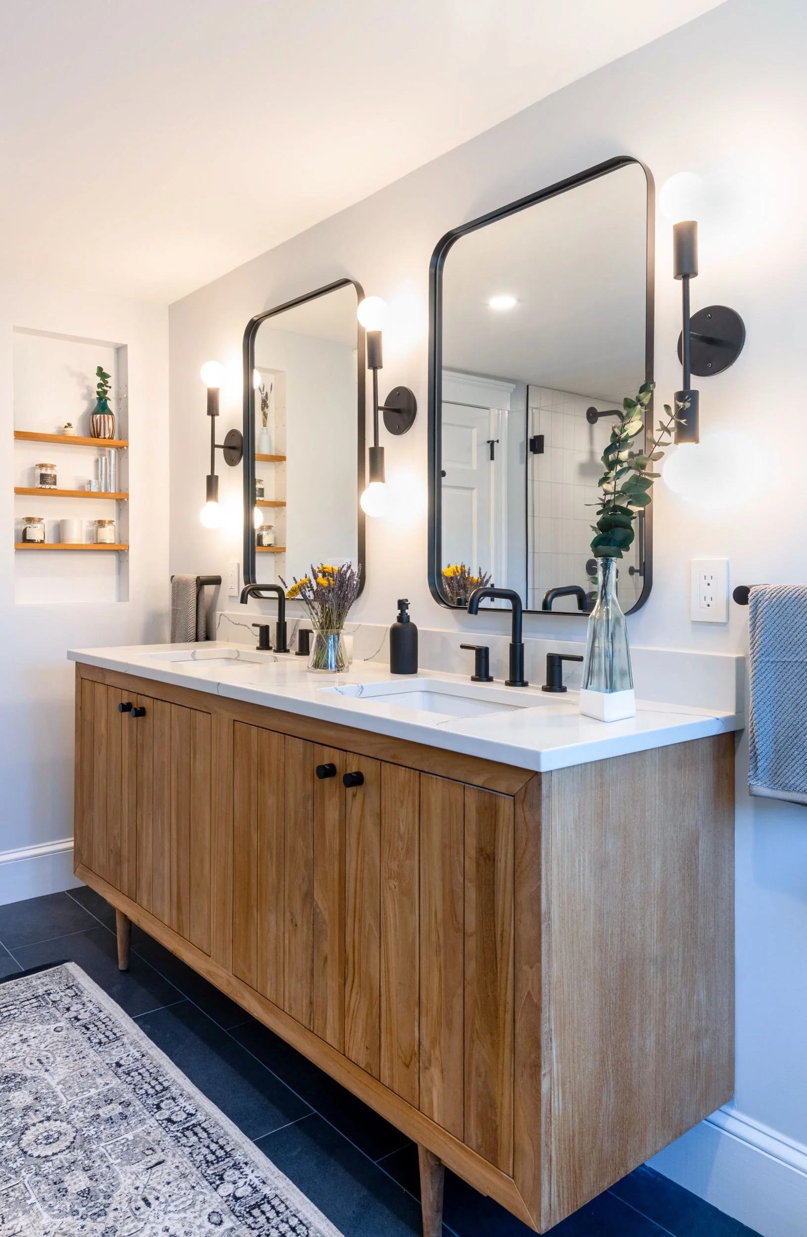 Teak vanity featuring double sinks and black fixtures with custom wall niche with teak shelves in this primary bathroom of a Medford, MA home.