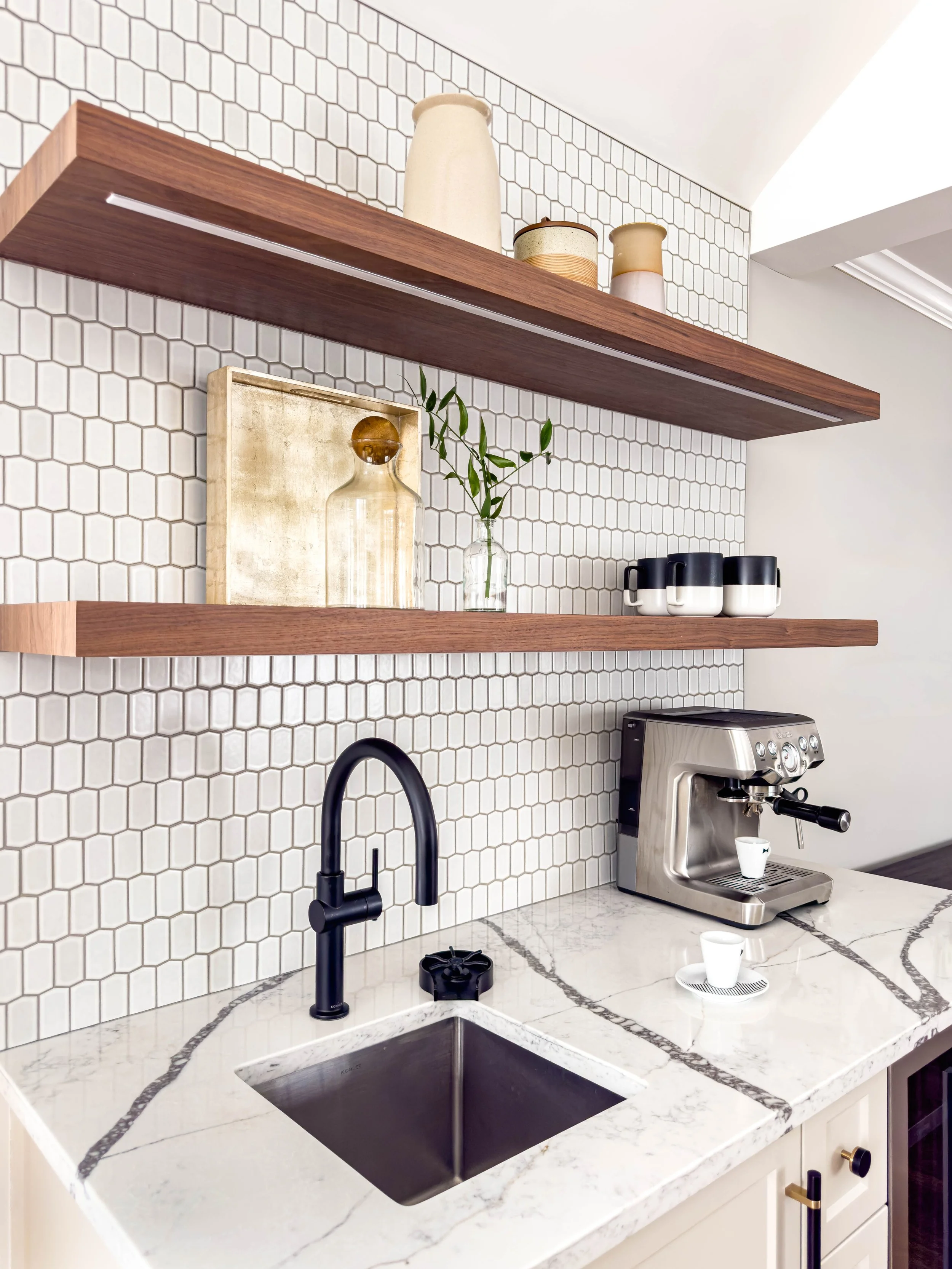 Kitchen coffee station detail featuring picket tile backsplash, floating walnut wood shelves with integrated LED lighting, and a marble-style quartz countertop with an undermount bar sink equipped with a glass rinser for high-end residential function