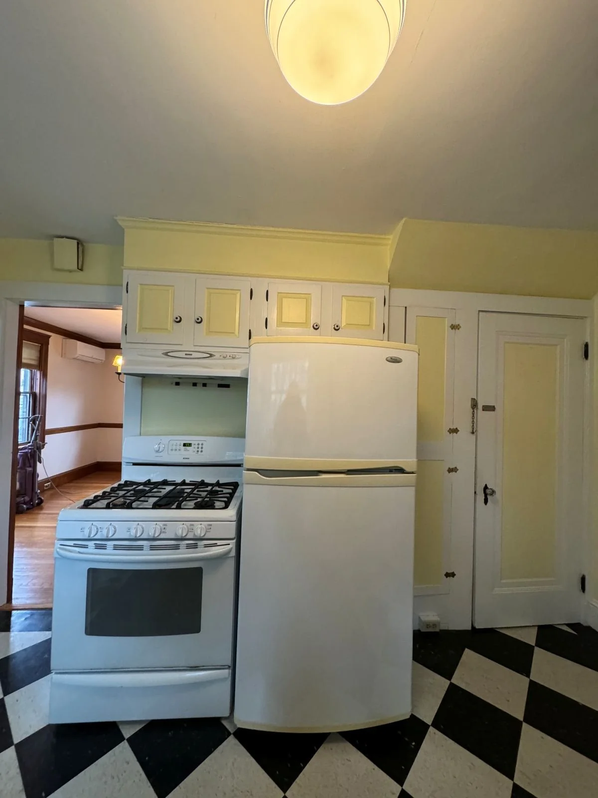 Existing kitchen range and refrigerator side by side surrounded by two-toned yellow and white cabinetry with view into dining room through cased opening