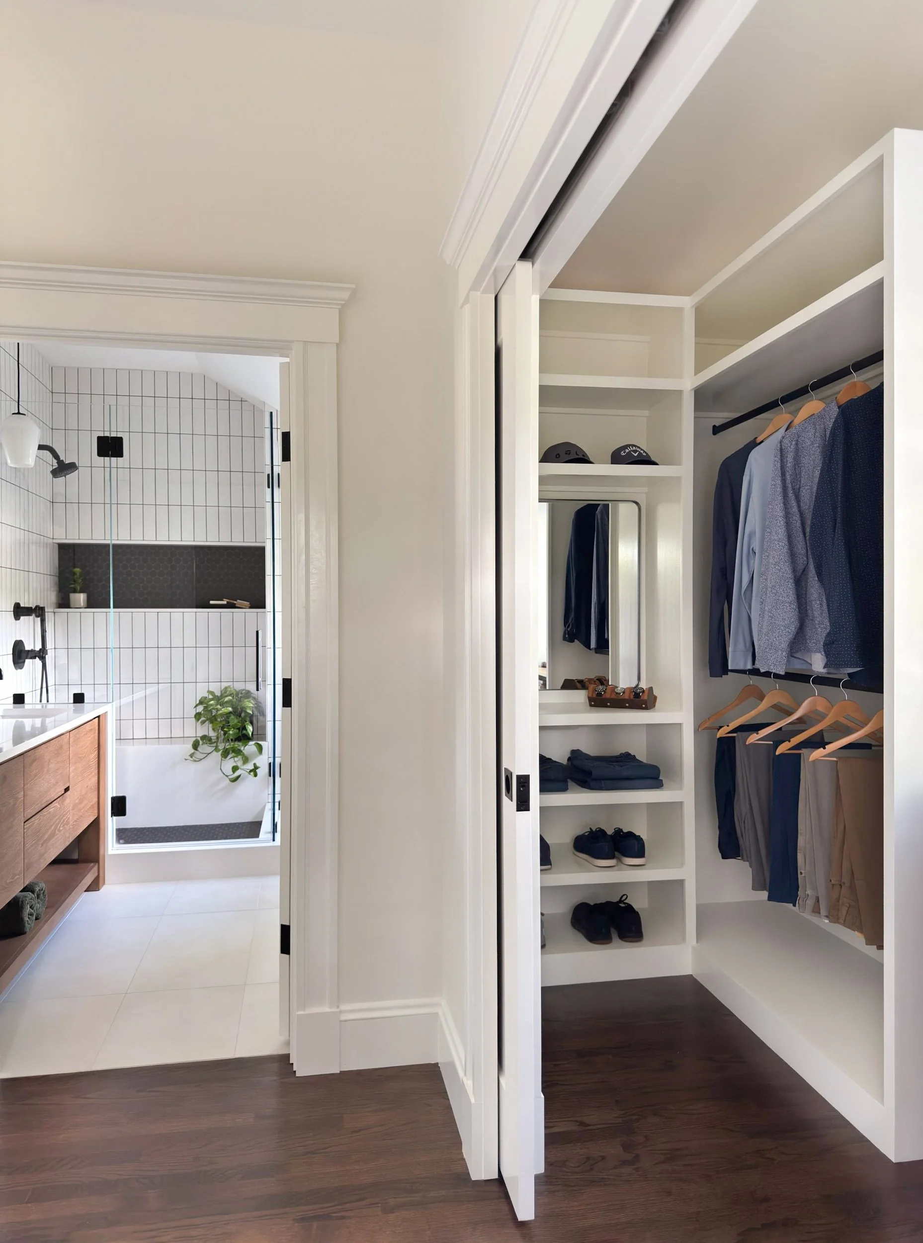 His side of a his and hers walk-in closet in a Dutch Colonial primary suite addition in Greater Boston viewed through pocket door showing custom built-in shelving with view beyond to primary bathroom.