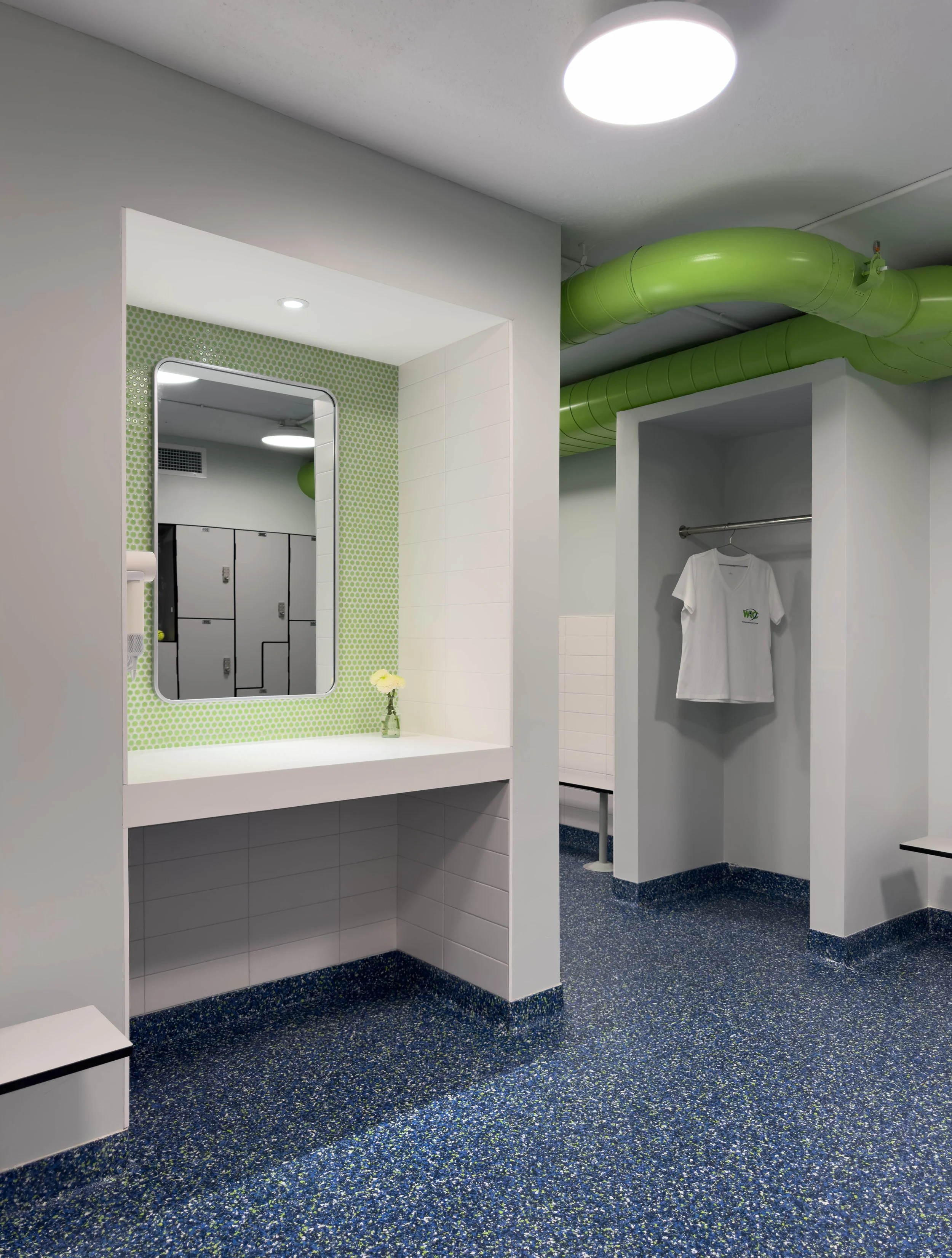 Completed commercial locker room renovation featuring green mosaic tile accent wall, integrated lockers, rounded mirror, and exposed green ductwork, designed by Tact & Grit Design Co.