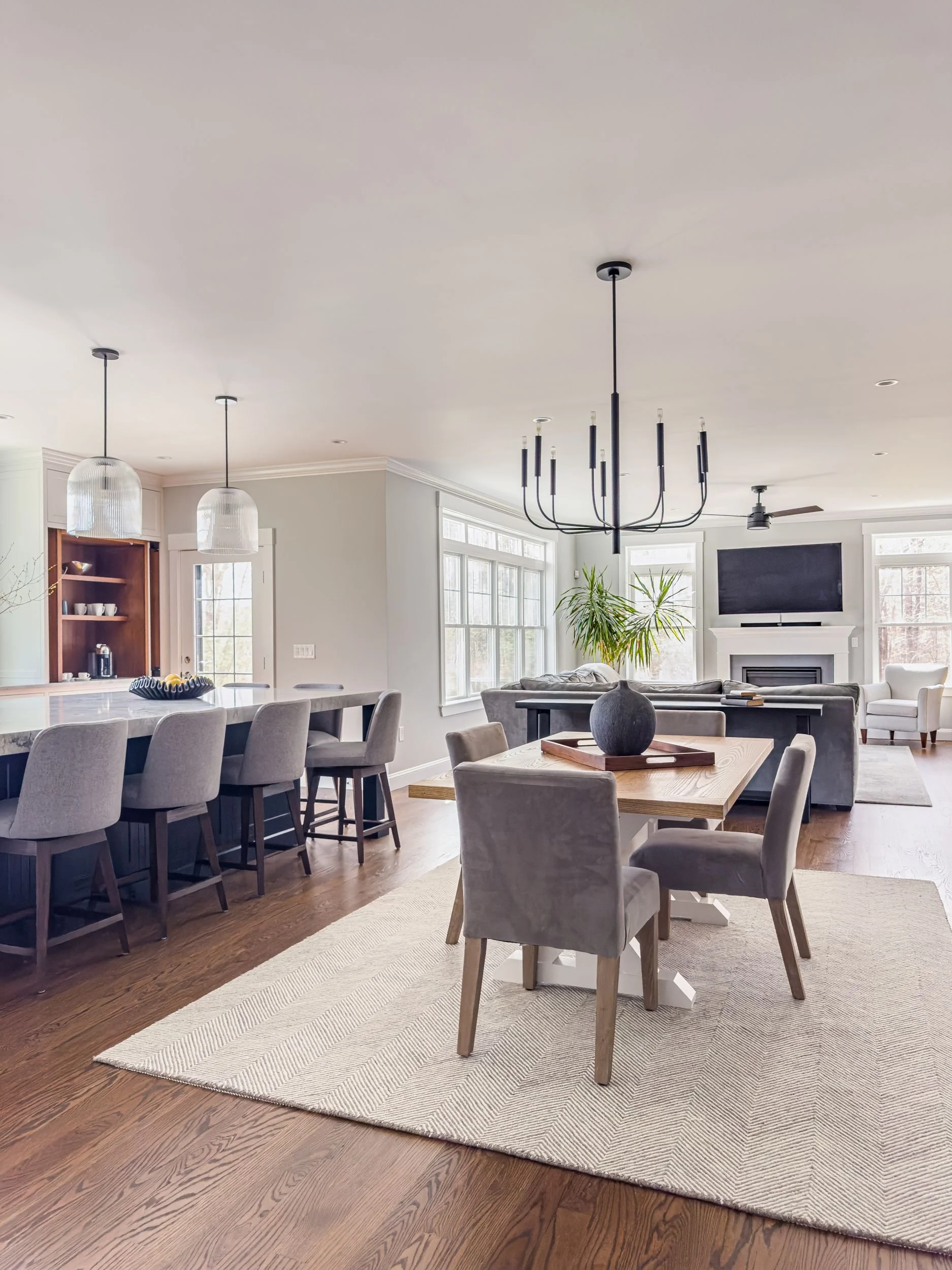Open-plan casual dining, kitchen, and living room with black chandelier, large island with seating, walnut dining table, and stained white oak accent cabinet in Hamilton MA home by Tact & Grit Design Co.
