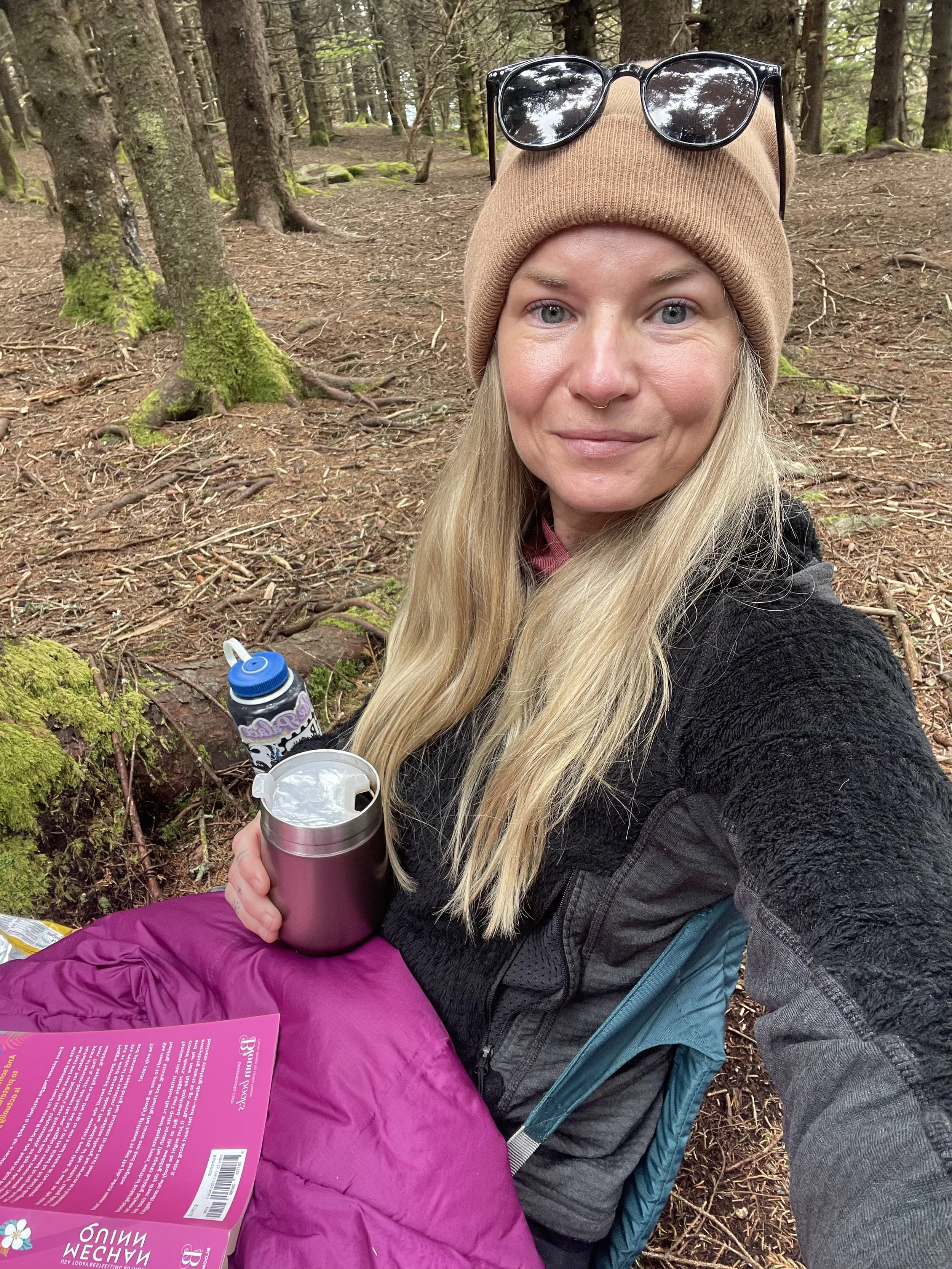 A woman with long blonde hair, wearing a tan beanie with sunglasses on top, is sitting in a forested area. She is holding a metallic tumbler and has a pink sleeping bag or blanket on her lap. She appears to be on a hiking or camping trip. Trees and m
