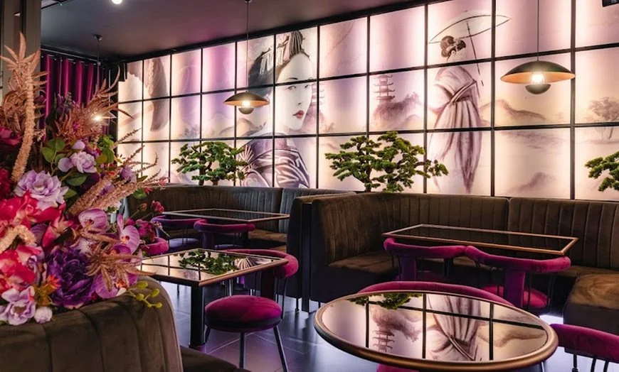 Elegant restaurant interior with black leather booths, pink velvet chairs, and small decorated tables. A large floral arrangement with pink, purple, and beige flowers is visible. The wall features backlit panels with traditional Japanese artwork and bonsai trees are placed on tables in front of the wall.
