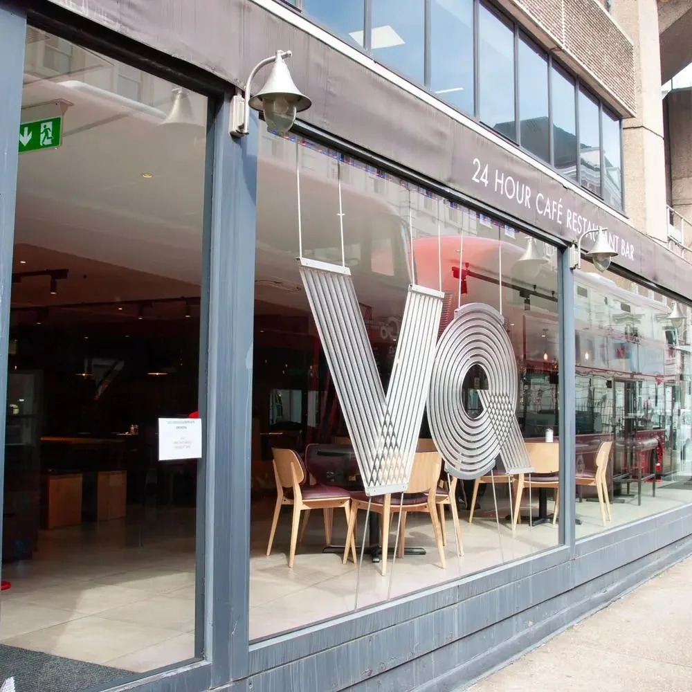 An outdoor restaurant patio at night with tables, chairs, umbrellas, and string lights. People are dining inside the restaurant, and a green neon sign is visible in the background. Trees and a building are also part of the scene.