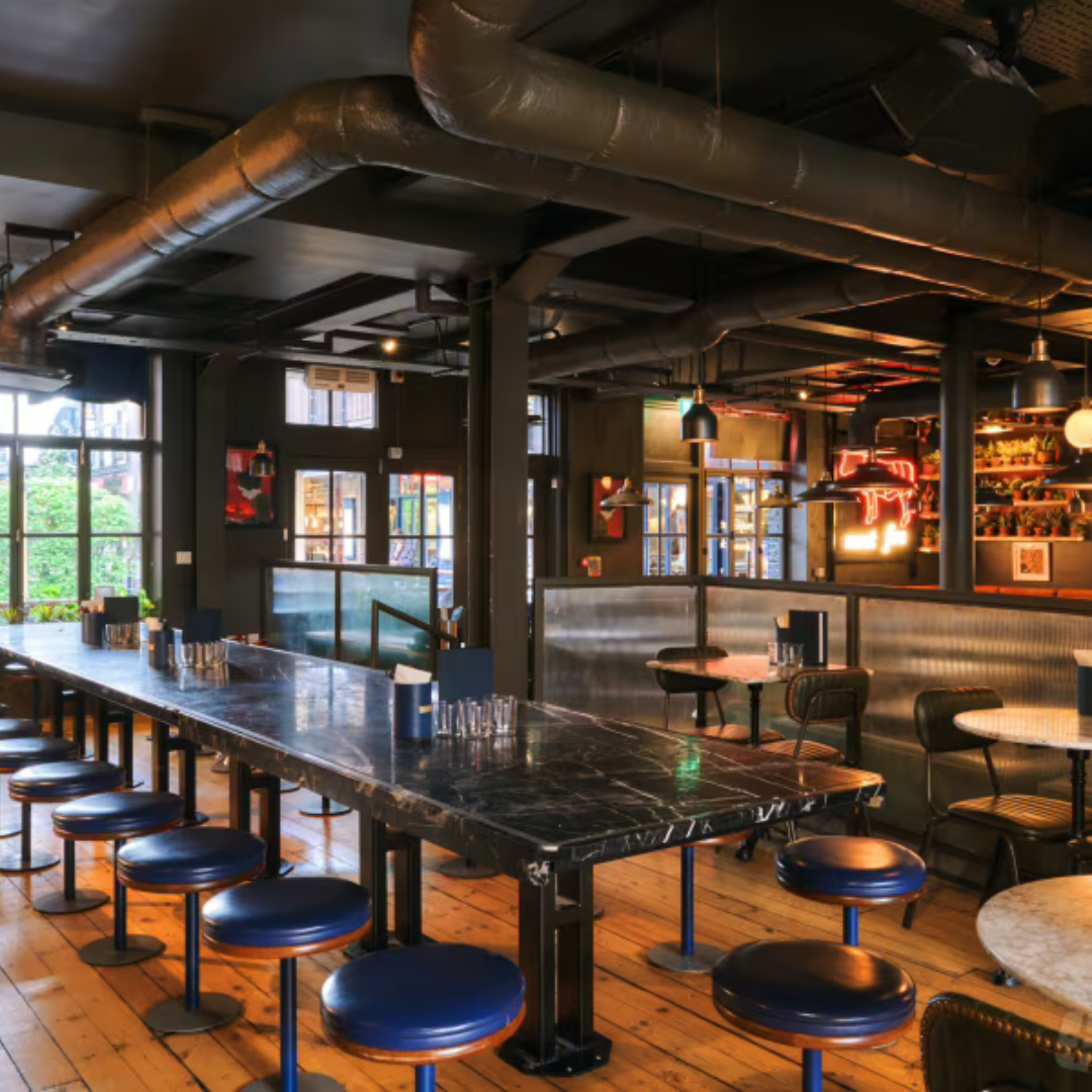 Interior of a modern café with tables, chairs, and bar seating, overlooking a bar area with bottles and glasses, illuminated by hanging pendant lights.