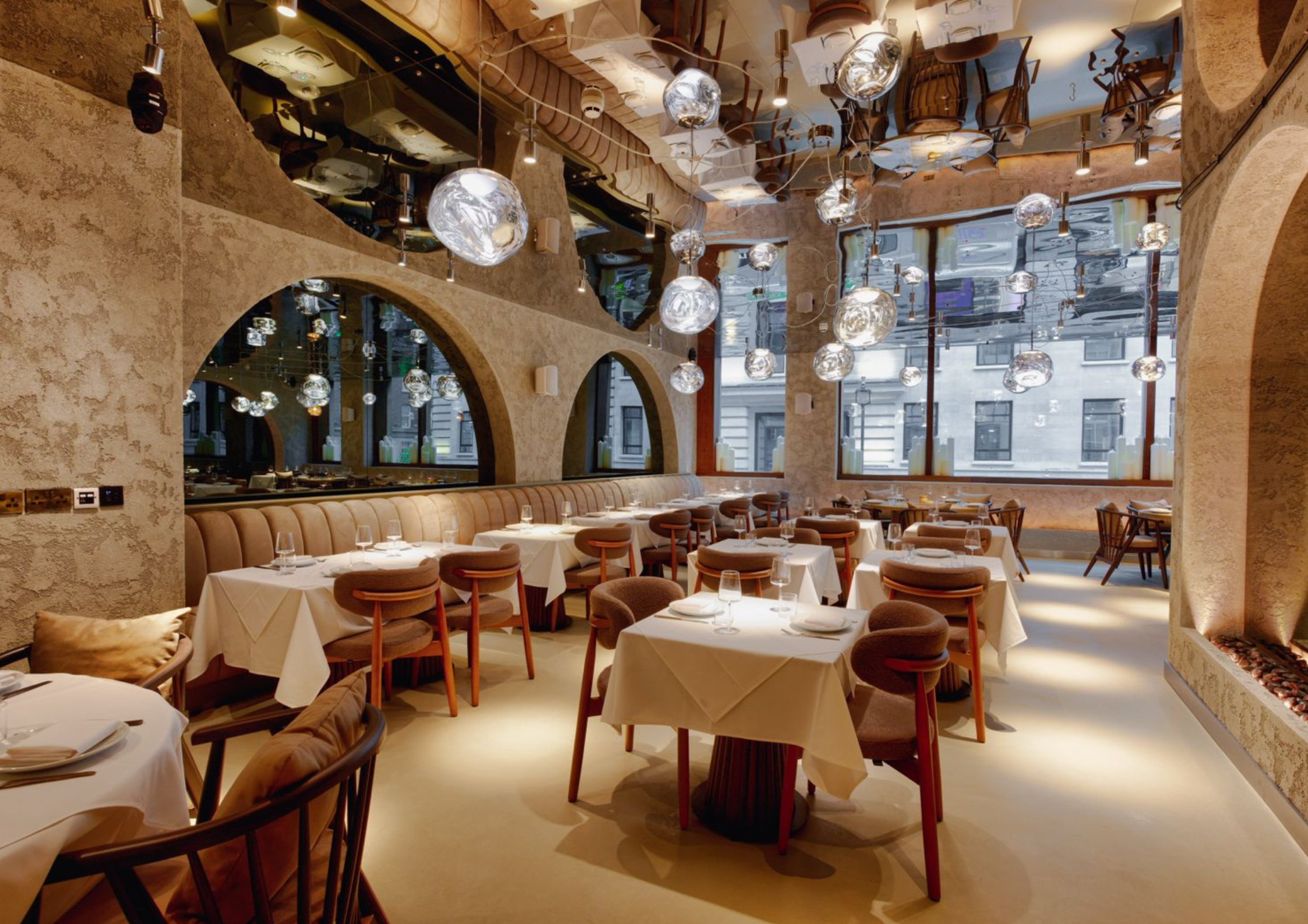A black two-story building with outdoor seating, potted plants, and signs advertising drinks and events, located next to a brick wall with a large shopping, eating, and drinking sign. The upper balcony features glass railing and greenery, and there's construction scaffolding on a tower in the background.