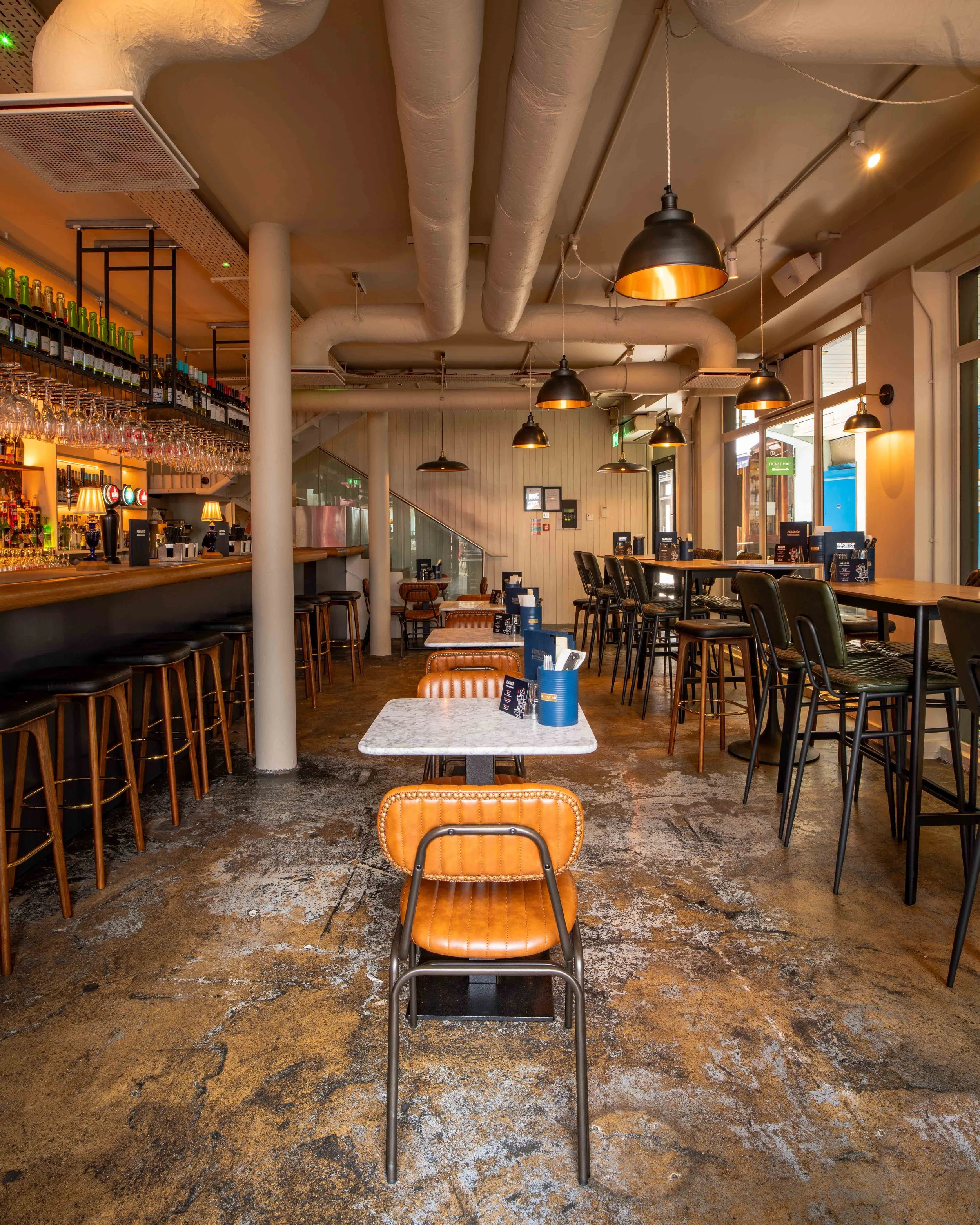 Interior of a modern café with tables, chairs, and bar seating, overlooking a bar area with bottles and glasses, illuminated by hanging pendant lights.