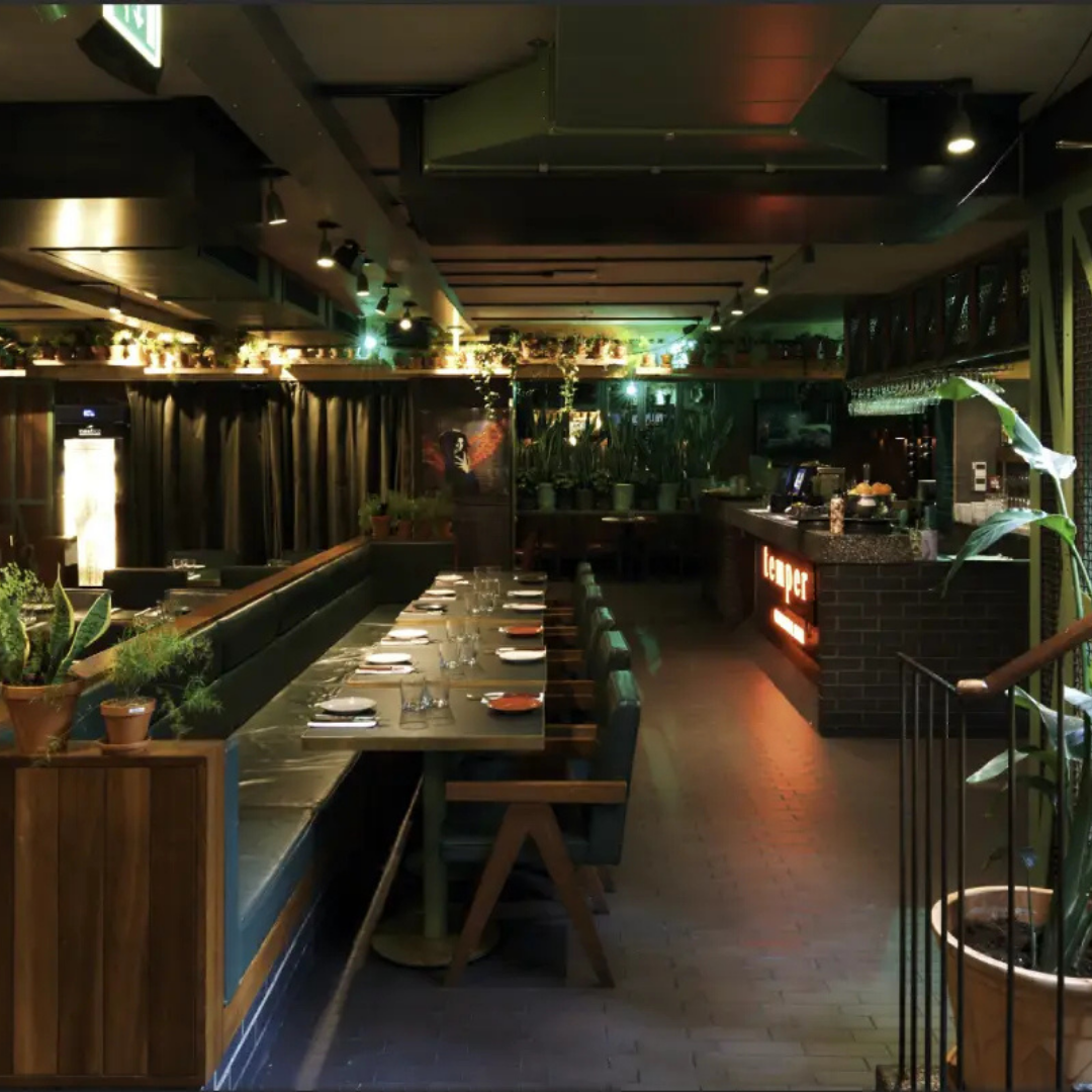 Empty restaurant with a central counter and red stools, modern decor, dim lighting, and dining tables set with glasses and utensils.