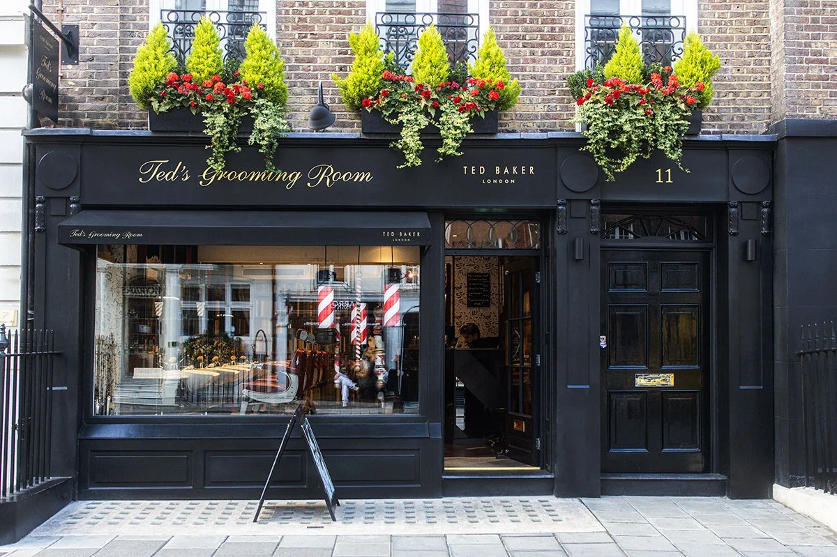 Exterior of Ted's Grooming Room, a black storefront with large window displaying barber poles and chairs. Potted yellow-green plants and red flowers decorate the upper part of the facade.