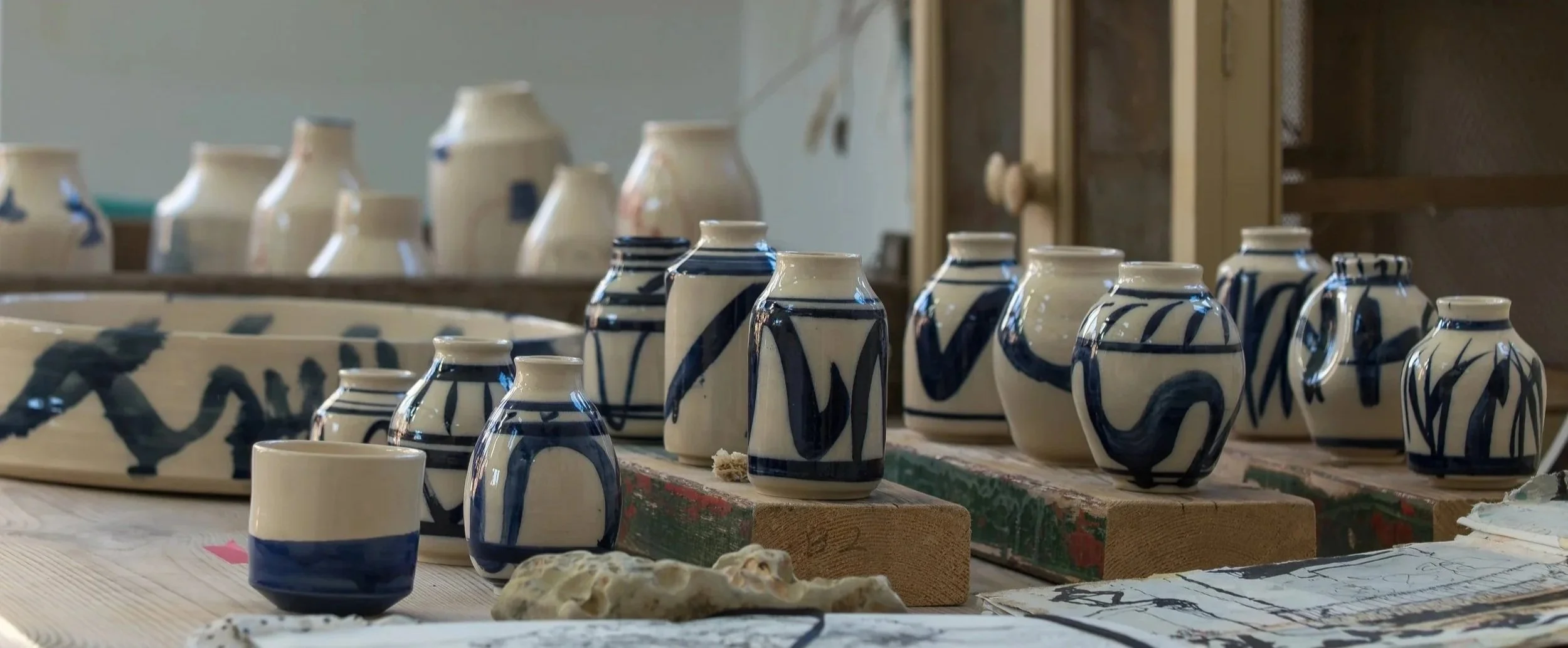 Collection of handmade pottery vases with blue and white abstract patterns on a wooden table, with some art supplies and sketches nearby.
