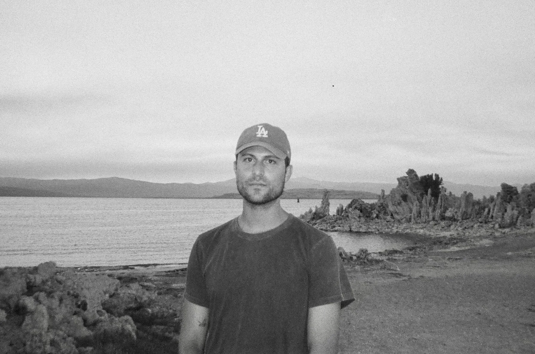 A man wearing a baseball cap and a t-shirt stands outdoors near a body of water, with a rocky shoreline and hills in the background, captured in black and white.