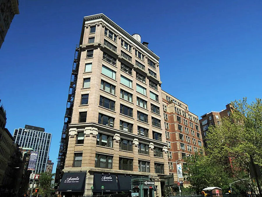 Tall historic city building with multiple windows and decorative architectural details, situated on a busy street with nearby buildings, trees, and a clear blue sky.