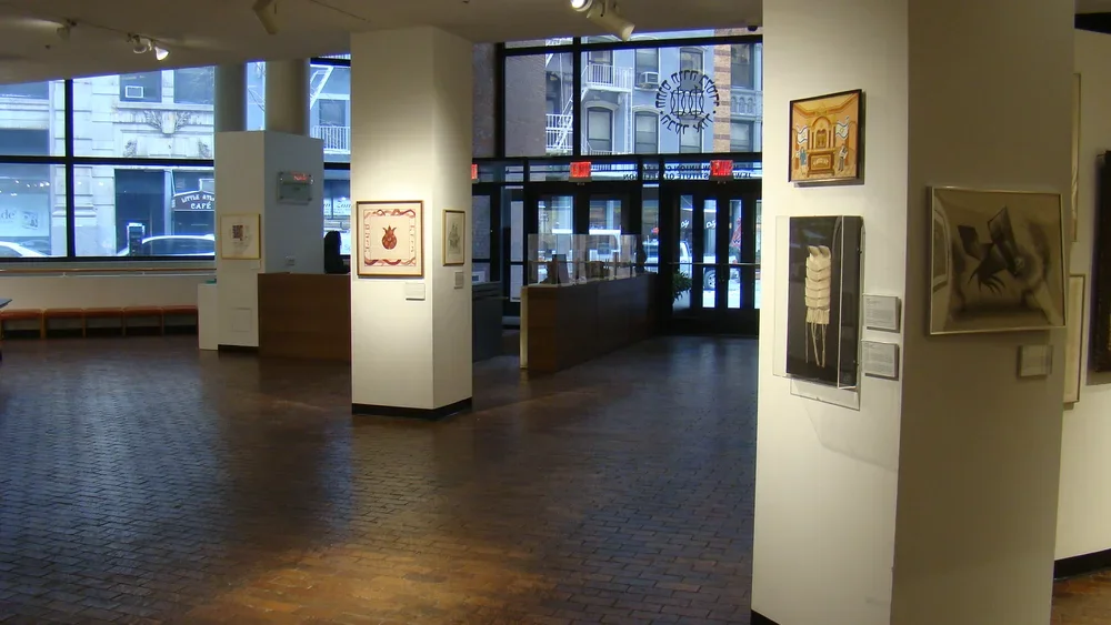 Interior of an art gallery with paintings displayed on white walls and a large window showing an urban street outside.