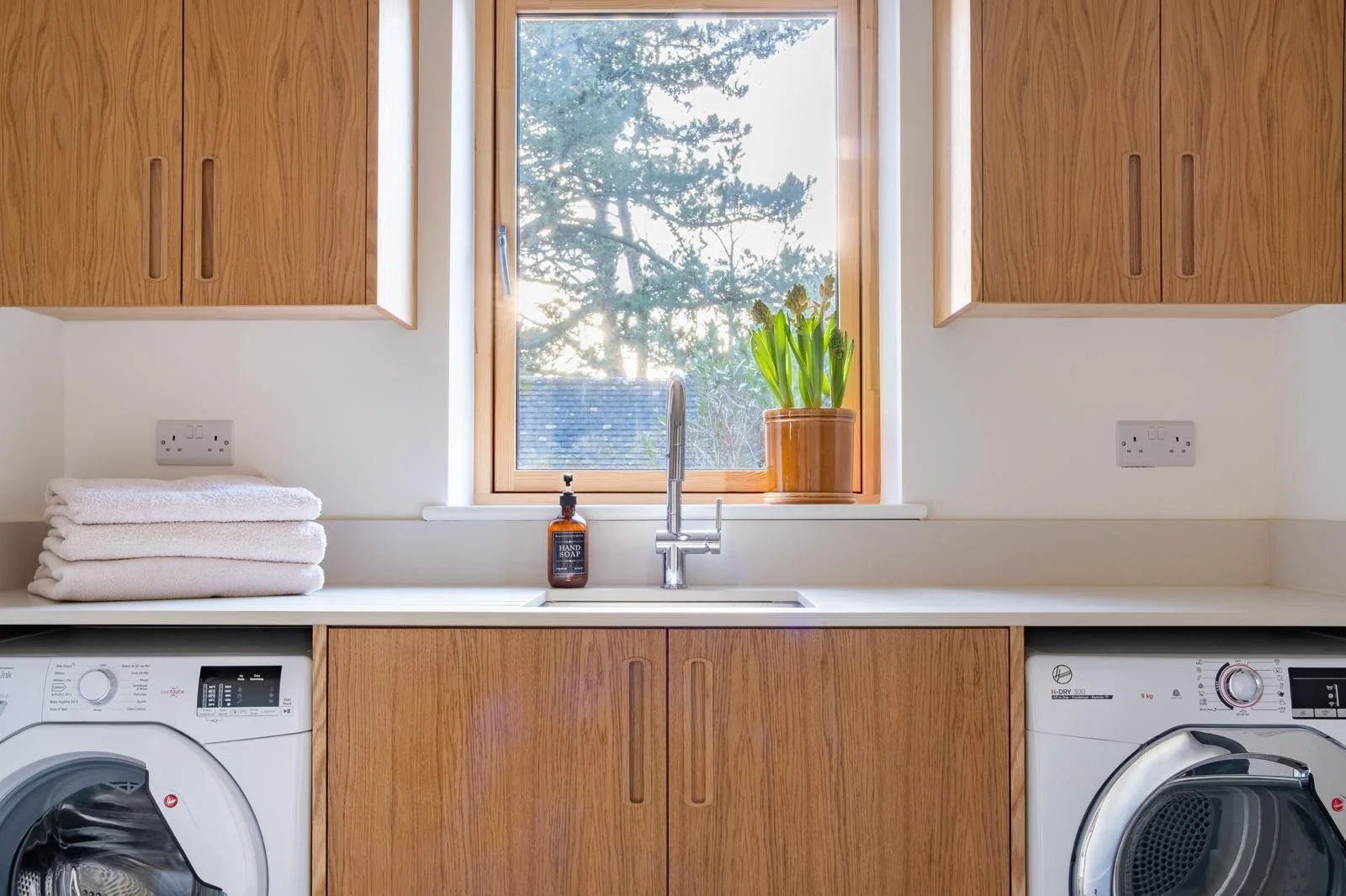 A laundry room with a window, wooden cabinets, a sink, a stack of towels, a bottle of hand soap, a potted plant, and two washing machines.