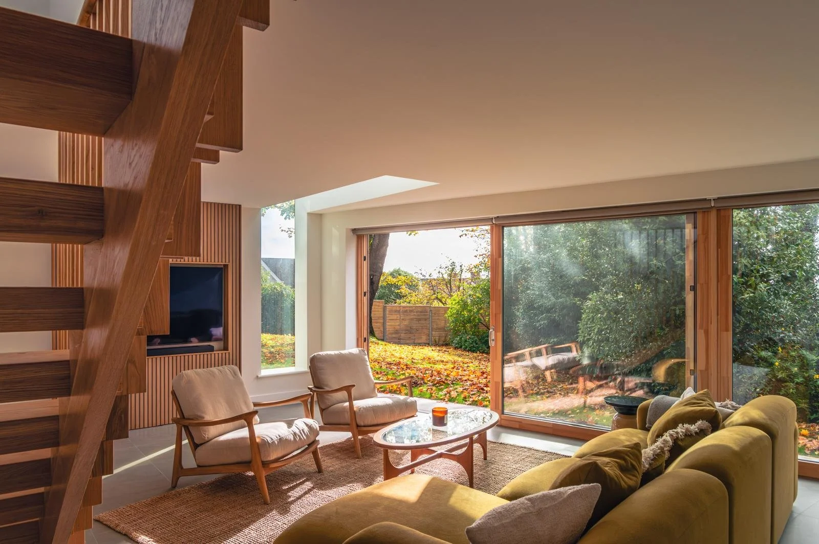 Modern living room with large glass sliding doors opening to a backyard with trees and fallen leaves, featuring a yellow sofa, two beige armchairs, a round glass coffee table, a textured rug, a wall-mounted TV, and wooden interior accents.