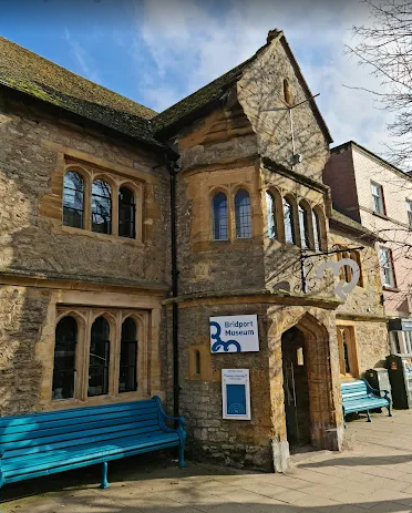 The exterior of the Bridport Museum, a historic stone building with arched windows, a gabled roof, and two blue benches outside.