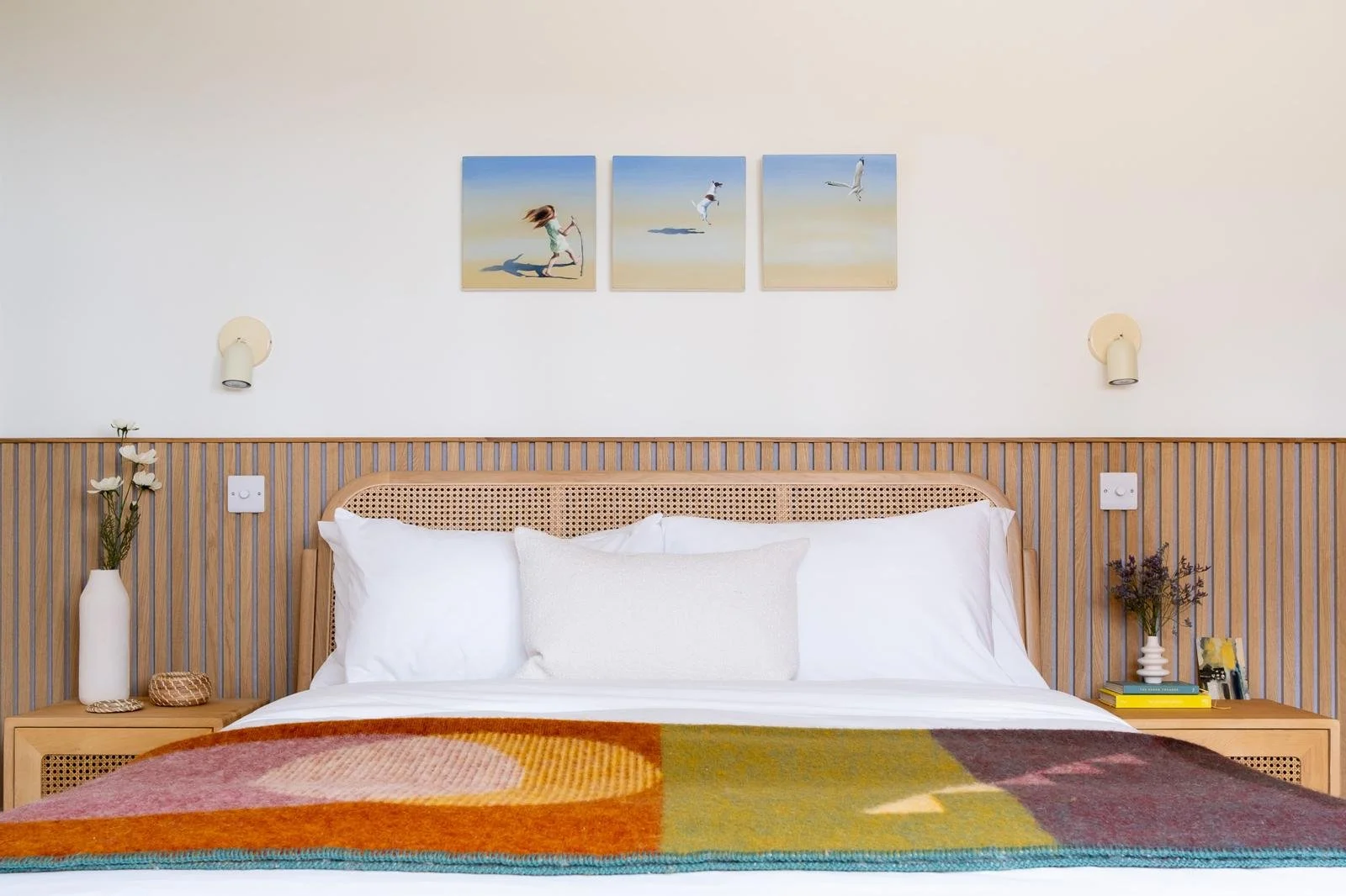 A neatly made bed with white pillows and a decorative pillow, a colorful blanket, two bedside tables with vases and flowers, and three seaside-themed art prints hanging on the wall above the bed.