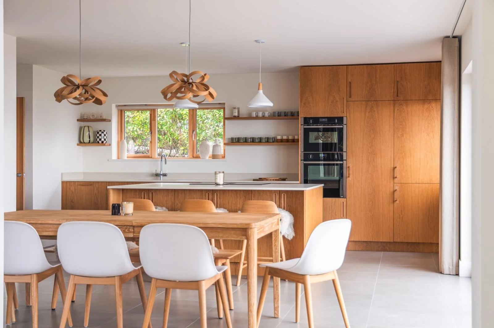 Modern kitchen with wooden cabinets, a dining table with white and wooden chairs, and decorative lighting fixtures.