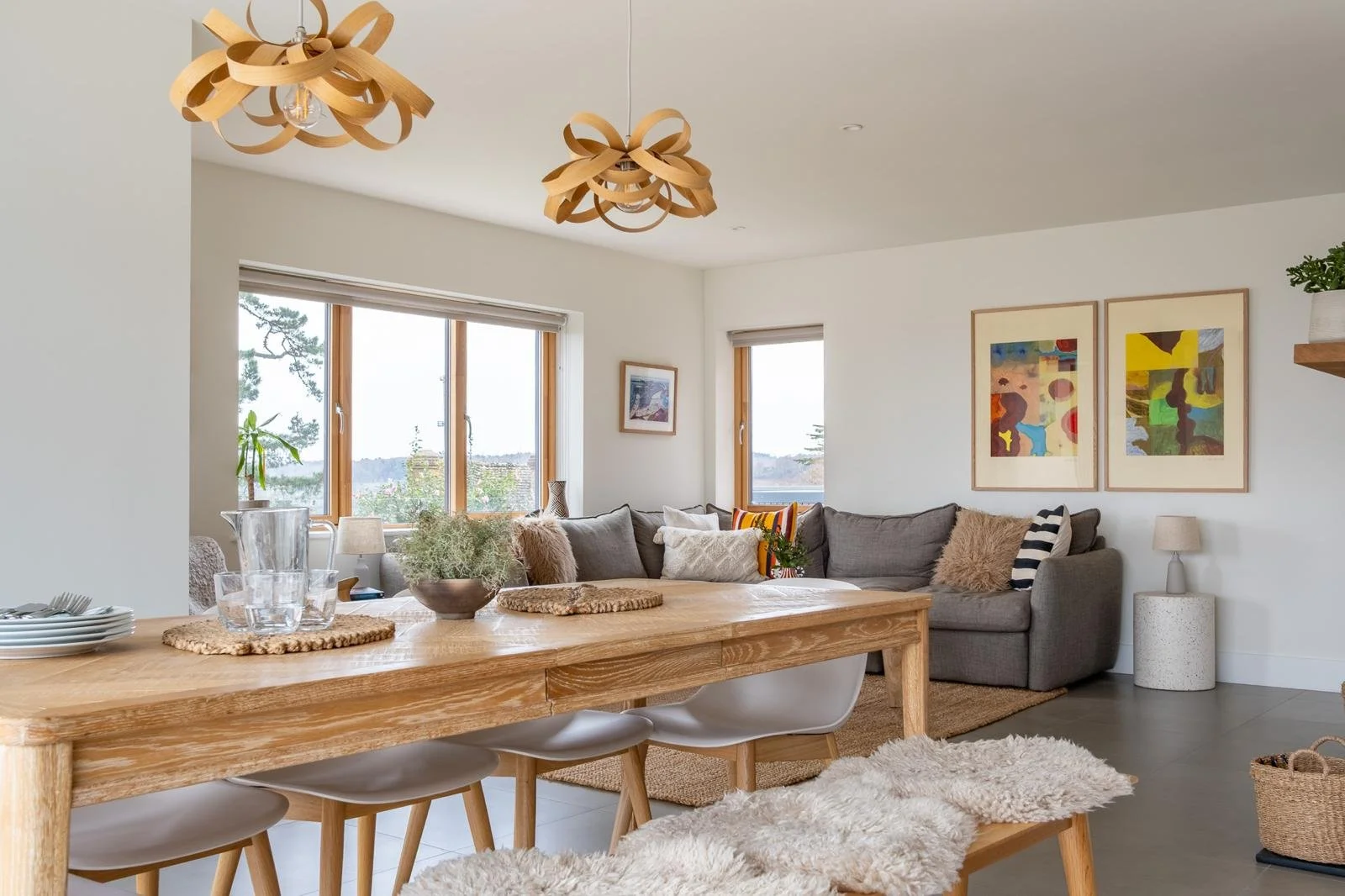 Living room with a cozy gray sectional sofa, colorful abstract art on the wall, and wooden ceiling lights. A large window with views of trees, and a wooden dining table with white chairs and a sheepskin throw.