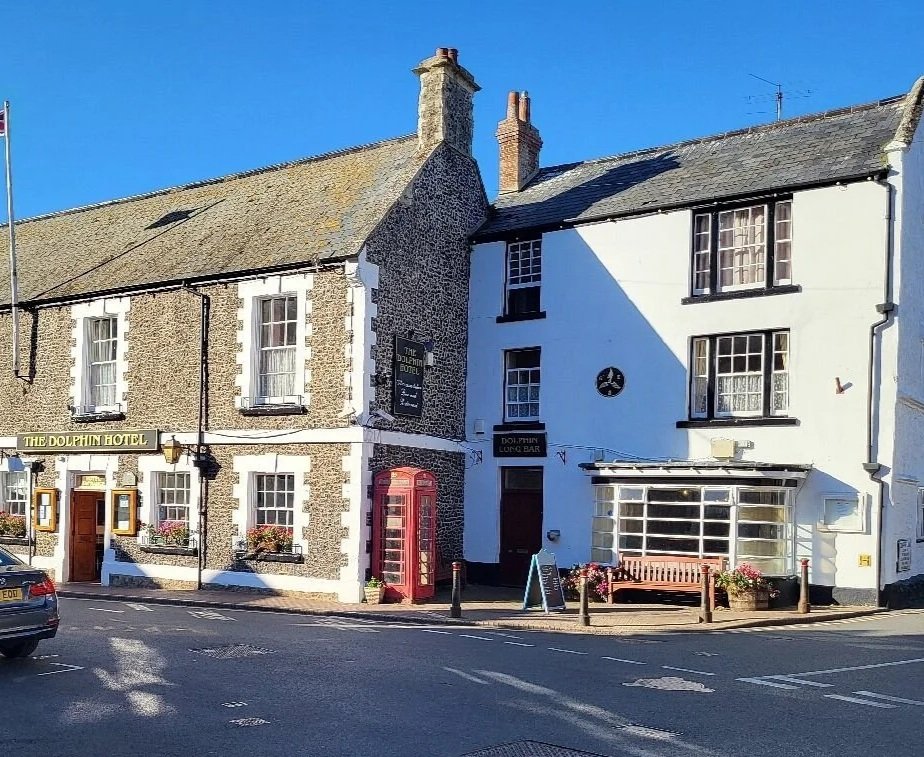 A street view featuring The Dolphin Hotel, a building with a half-timbered and white wall facade, a red telephone booth, a pink bench, and flower pots under a clear blue sky.