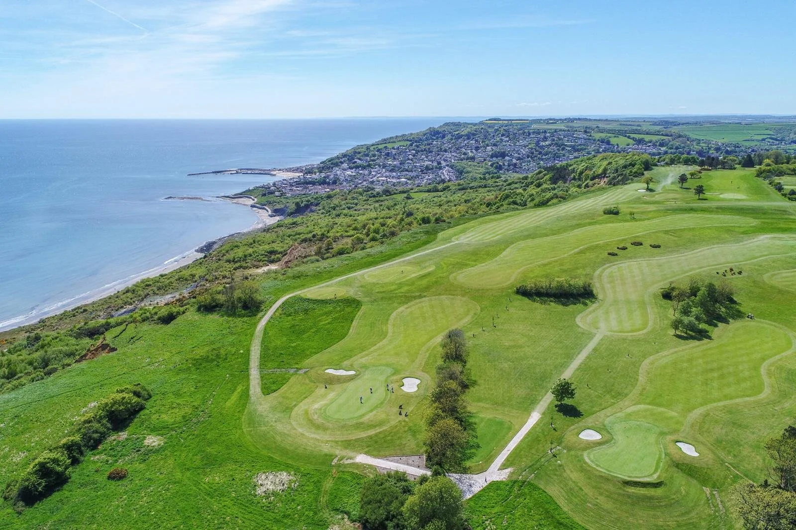 An aerial view of a golf course situated along the coastline, with lush green fairways, sand bunkers, scattered trees, and the ocean in the background.