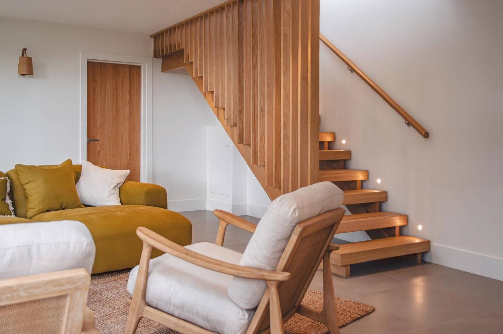 Living room with wooden staircase, white walls, beige and mustard-colored furniture, and warm lighting.