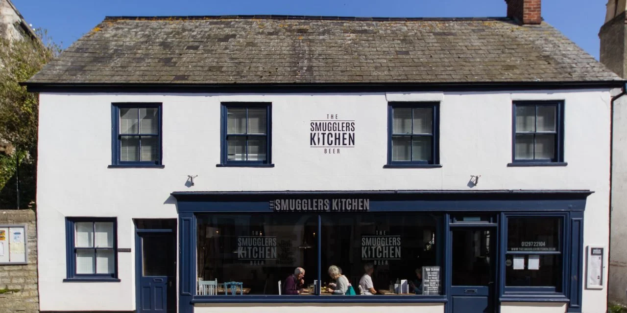 White two-story building with black window frames and a black awning; signage displays 'The Smugglers Kitchen Beer.' Inside, people are sitting at tables.
