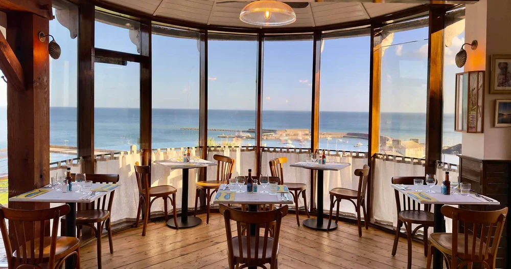 Dinner tables with place settings, in a restaurant with large bay windows, overlooking the ocean and a harbor.