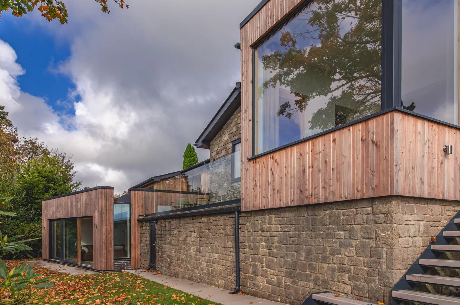 Exterior view of a modern house with a stone foundation, wooden siding, large glass windows, and black trim, surrounded by a lawn with fallen leaves during daytime with cloudy sky.