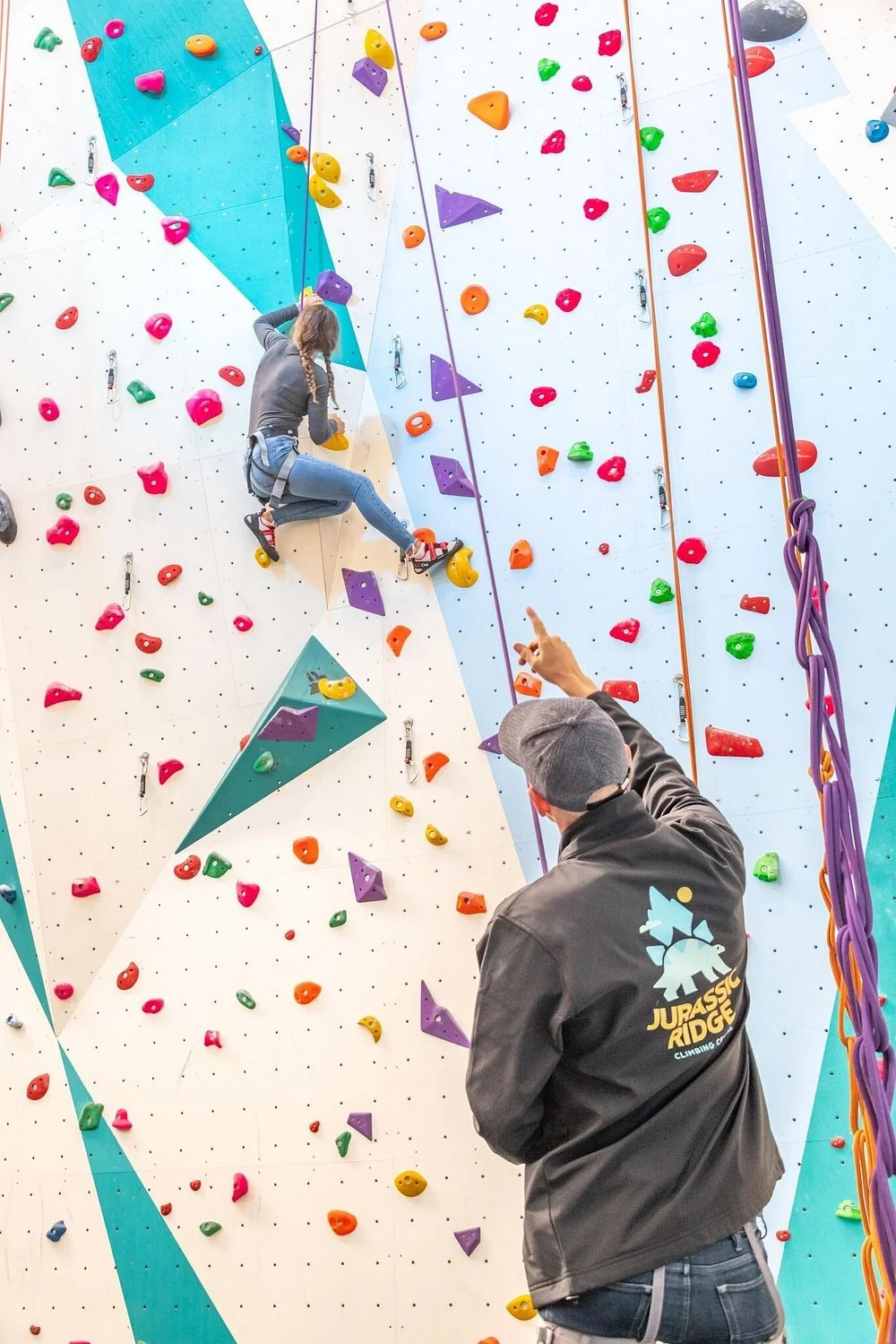 A woman is climbing an indoor rock climbing wall with colorful holds, while a man in a dark jacket and cap offers guidance or instructions. The woman is wearing a harness and climbing shoes.