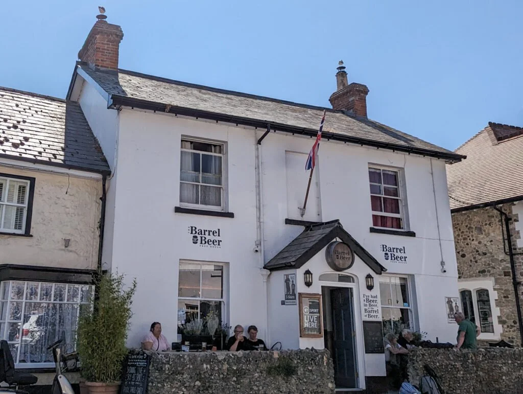 Outside view of the Barrel & Beer pub, a white two-story building with black trim, a small porch with a sign, and people sitting outside.