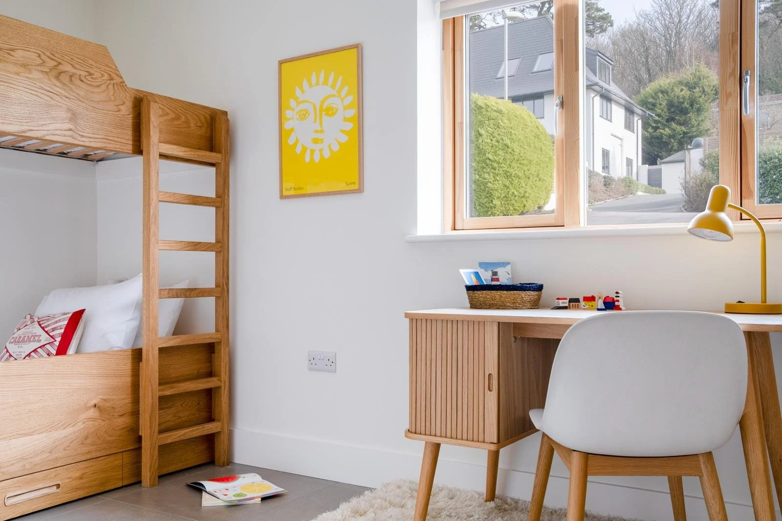 Children's bedroom with wooden bunk bed, white walls, a yellow framed poster, a desk with a yellow lamp, and a window showing a residential neighborhood outside.