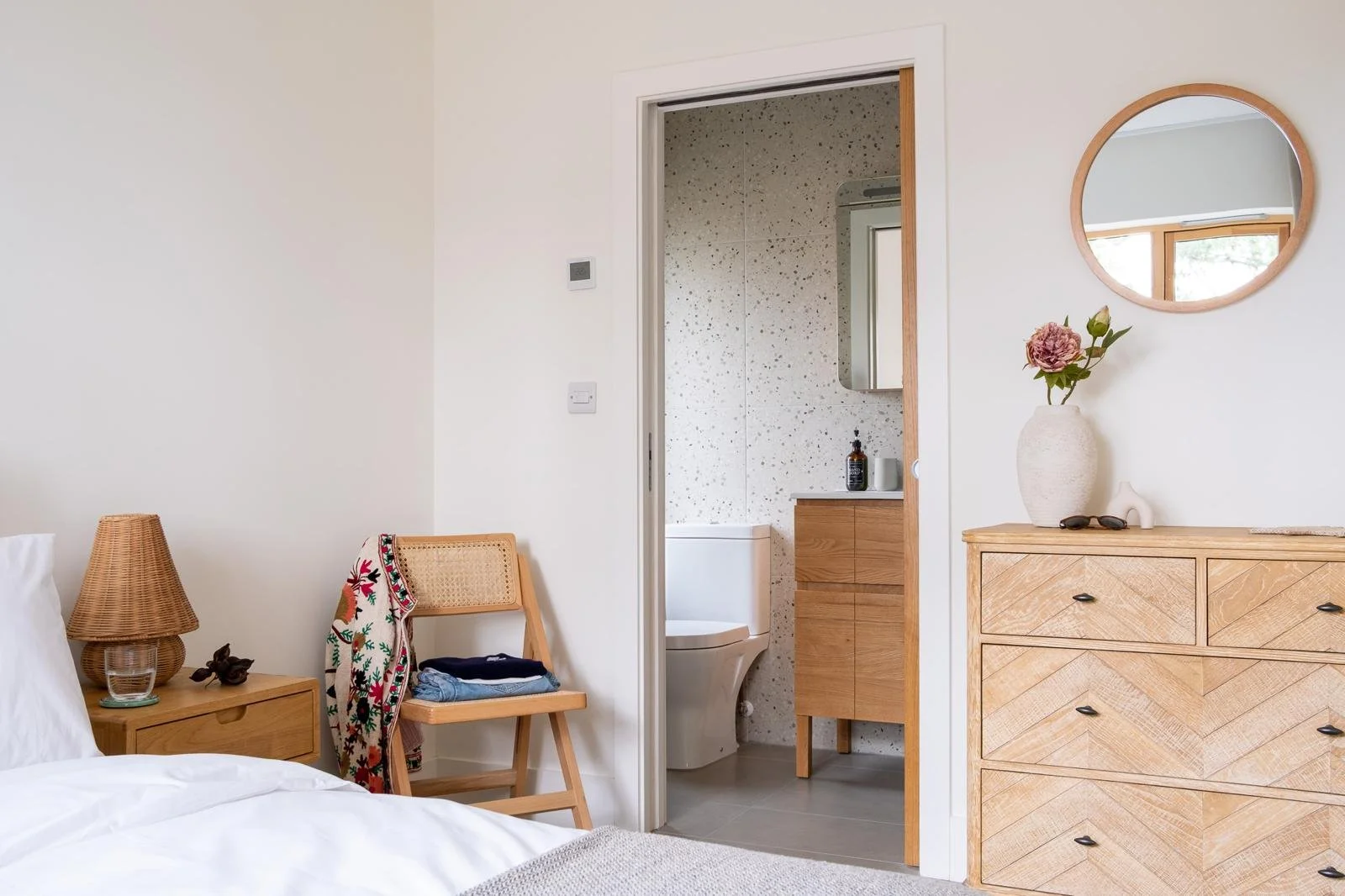 A bedroom with neutral tones, wooden furniture, a nightstand with a wicker lamp, a woven chair with a patterned cloth and clothing, a dresser with a vase with pink flowers, a mirror, and a doorway leading to a bathroom with terrazzo walls, a toilet, 