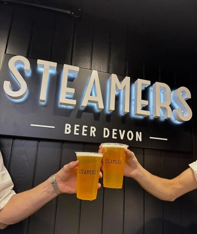 Two people holding cups of beer in front of a sign that says 'STEAMERS' and 'BEER DEVON' in a dimly lit bar or brewery.