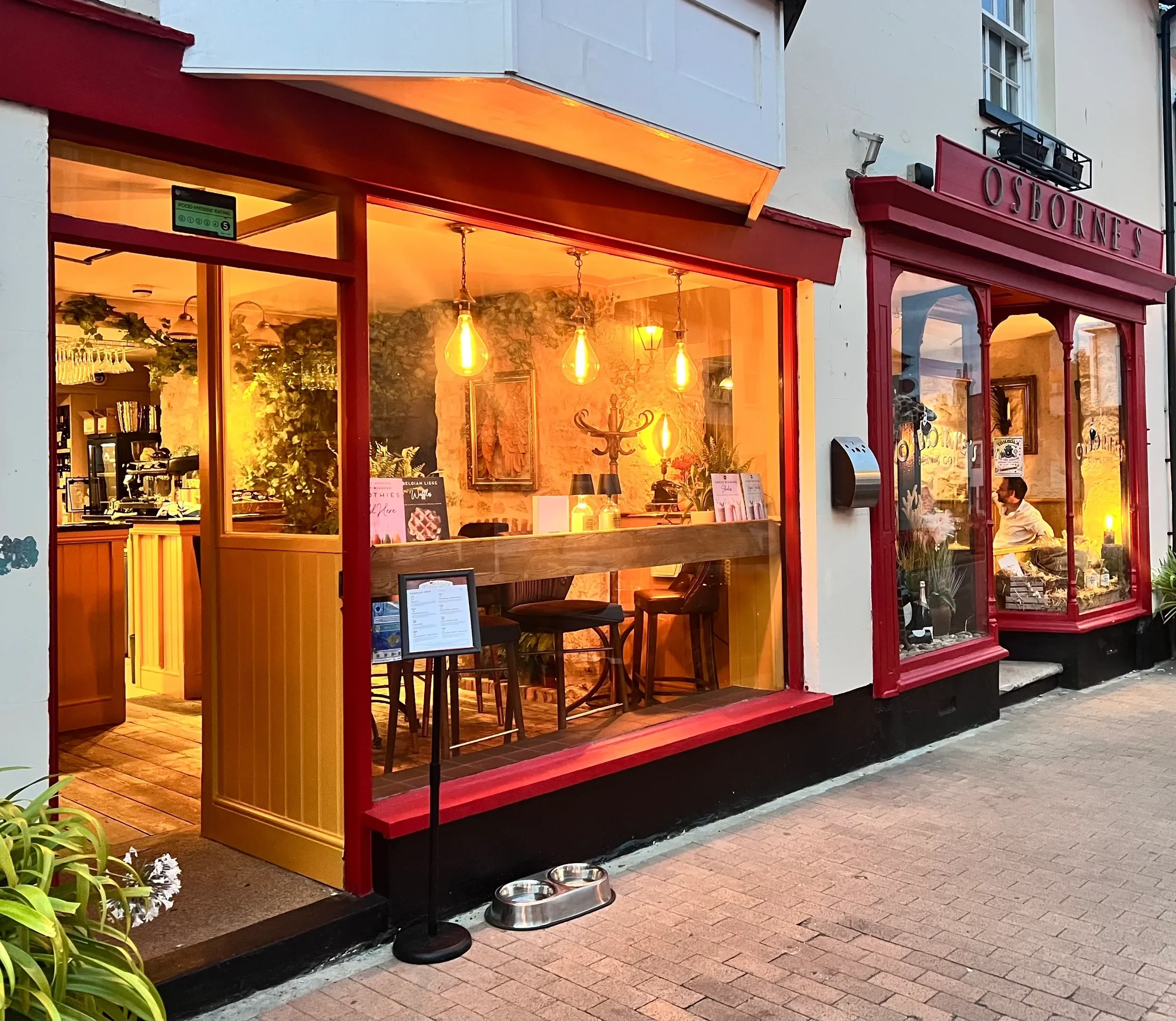 Exterior of a cozy restaurant with large windows, warm lighting, and a red and white facade named Osbourne's, with small plants outside.