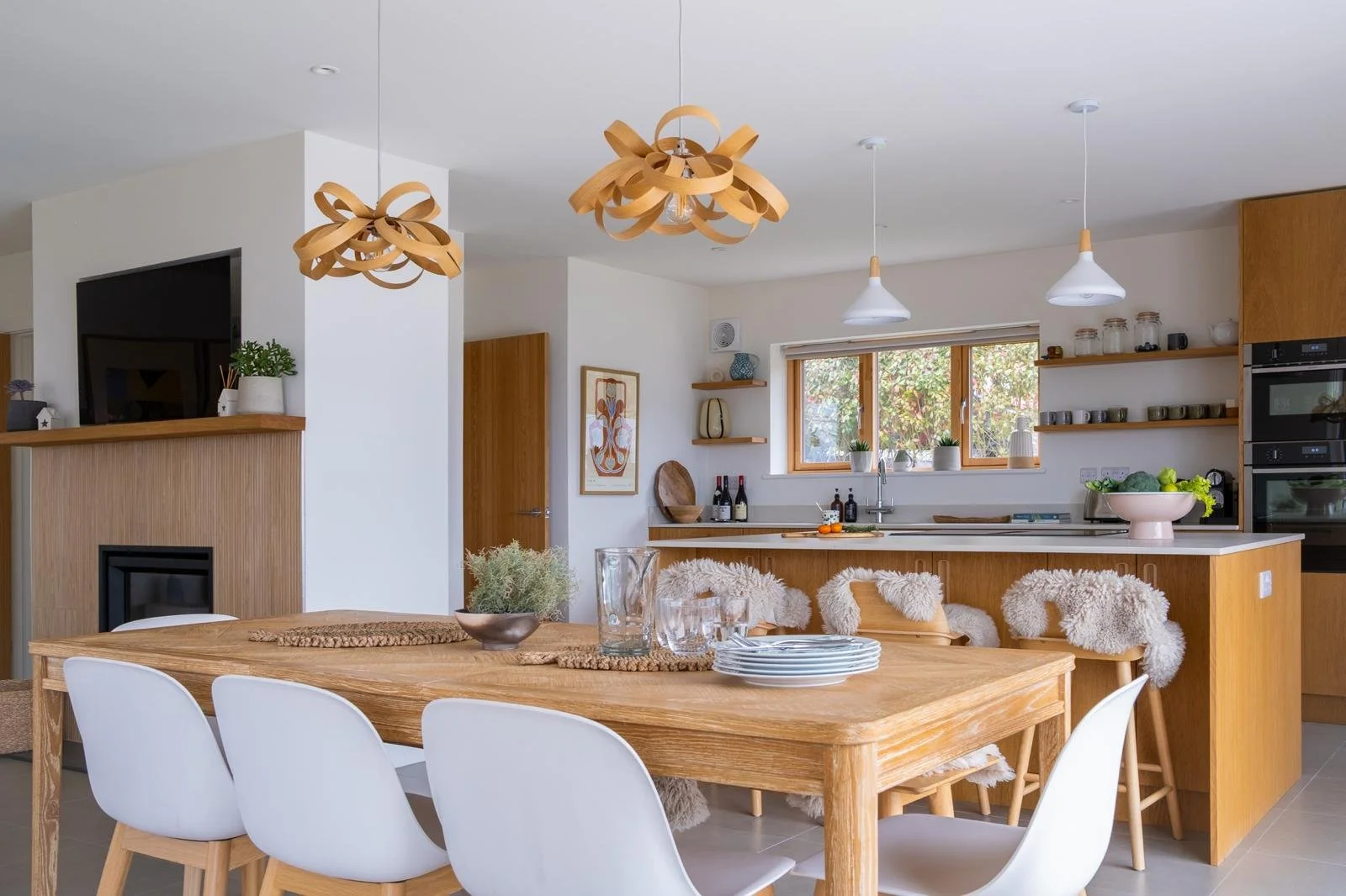 Modern kitchen with wooden accents, white walls, hanging light fixtures, and a dining table with white chairs.