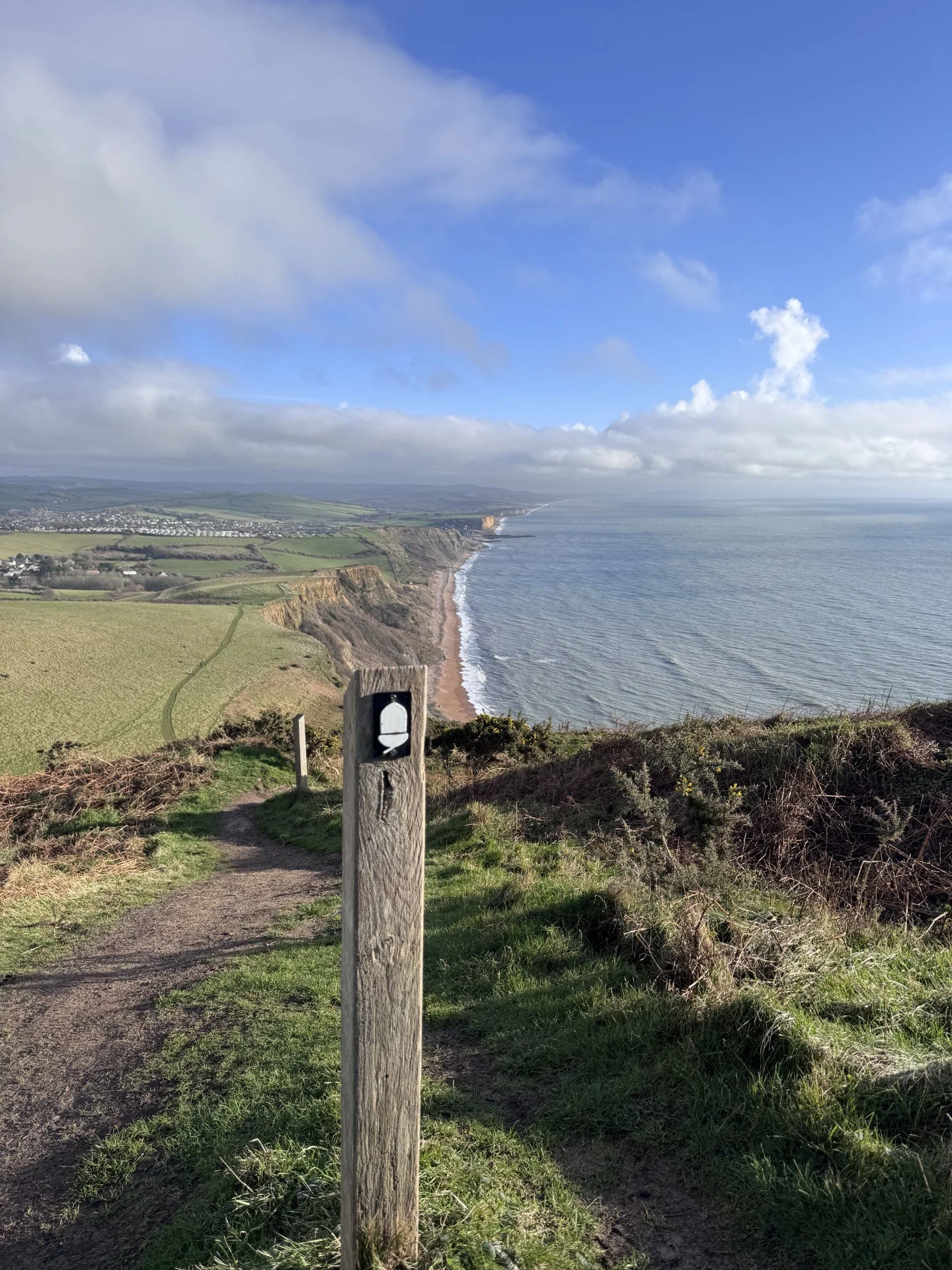 A scenic coastal view from a cliff with a dirt trail and a wooden marker post in the foreground. The ocean and a distant shoreline are visible under a partly cloudy sky.