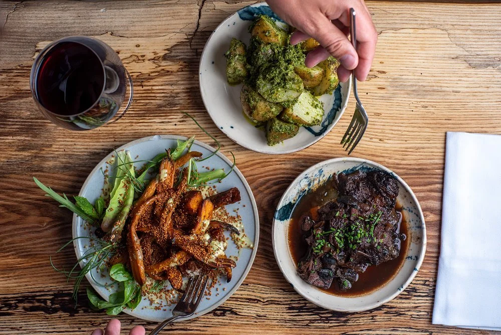 A top-down view of a wooden table with three plates of food and a glass of red wine. The plates contain roasted vegetables topped with herbs, a beef stew in dark sauce garnished with green herbs, and a side of seasoned potatoes. Hands are seen holding a fork and a spoon.