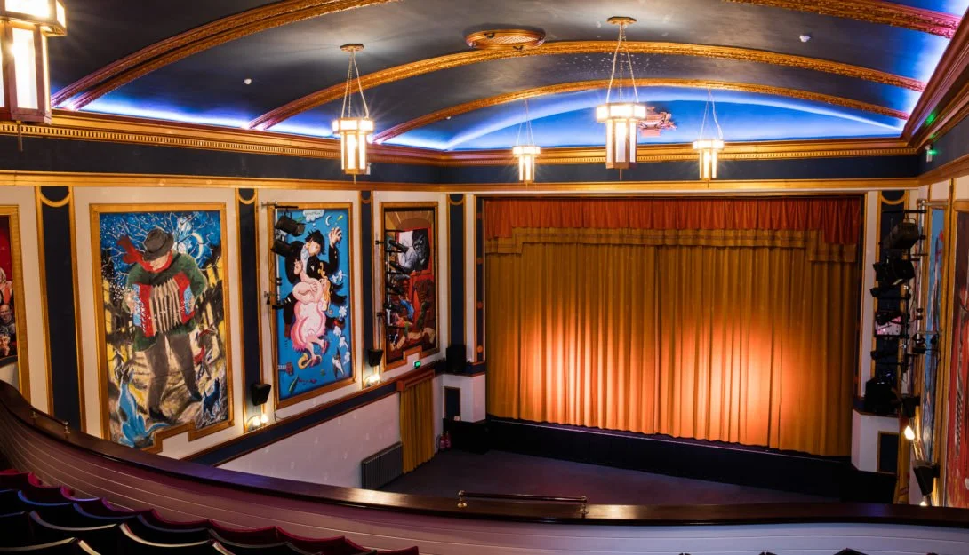Interior of a theater with a stage, golden curtains, colorful wall paintings, ornate ceiling, and stage lights.