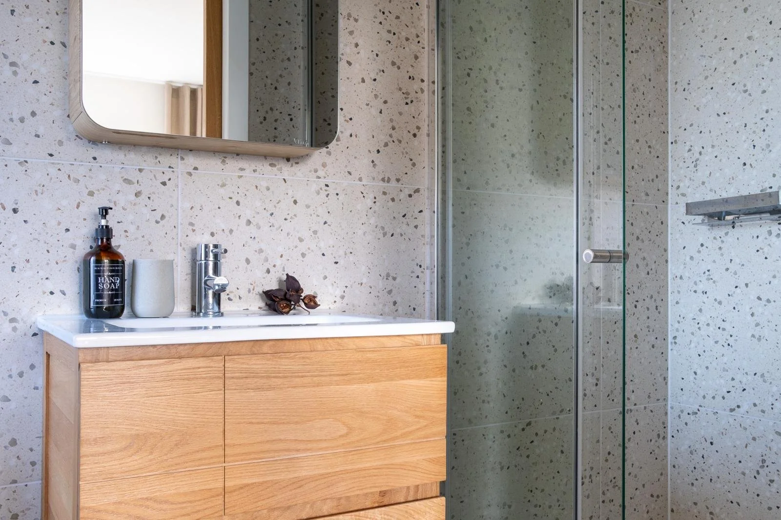 Modern bathroom with a wooden vanity, a mirror, a soap dispenser, and a black soap bottle. A glass shower door with a metal handle, and a speckled tile wall.