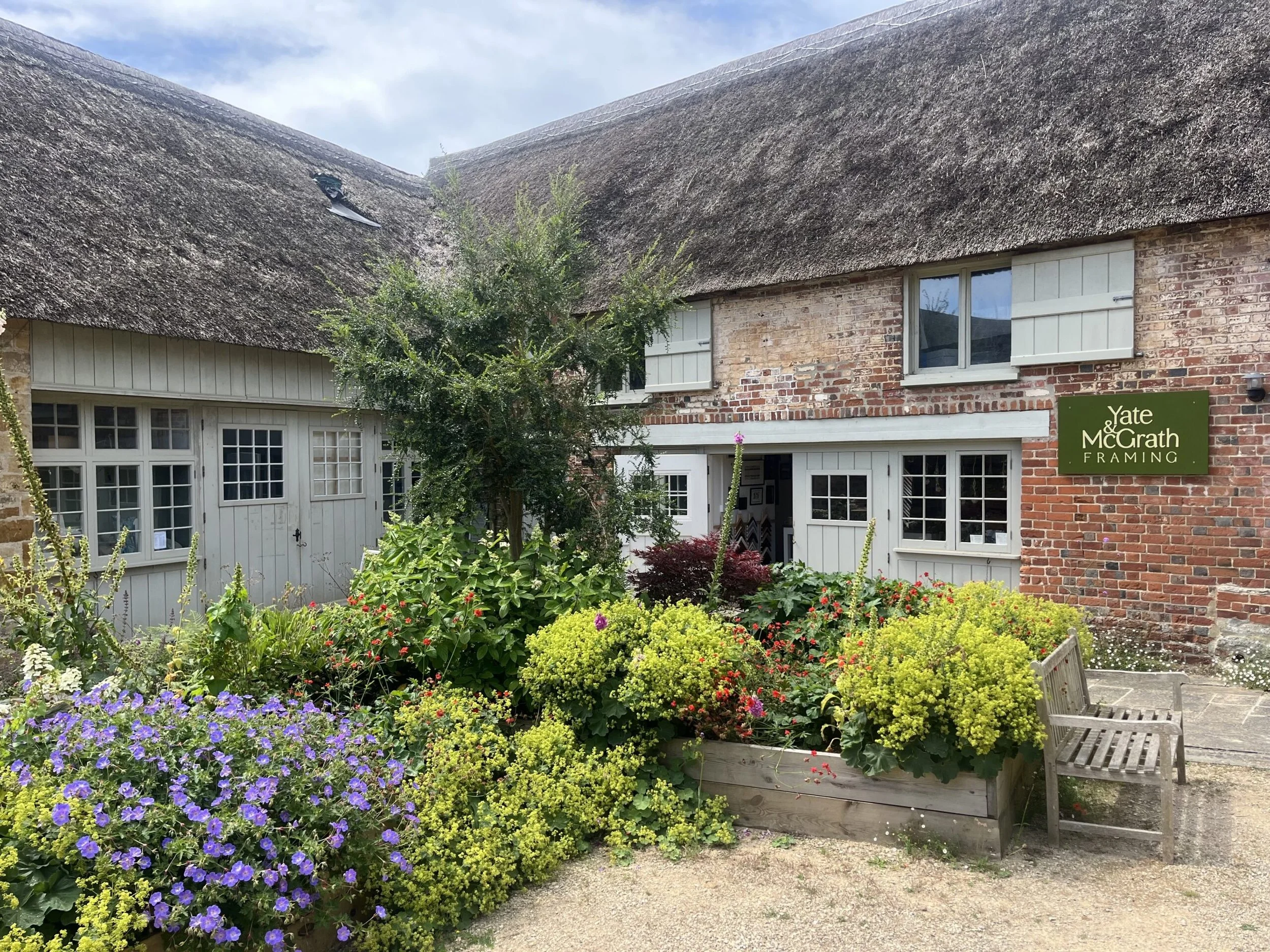 A garden with colorful flowers in front of a rustic brick house with white window shutters and a green sign that reads "Yate McGrath Framing." The house has a thatched roof and a small wooden bench nearby.