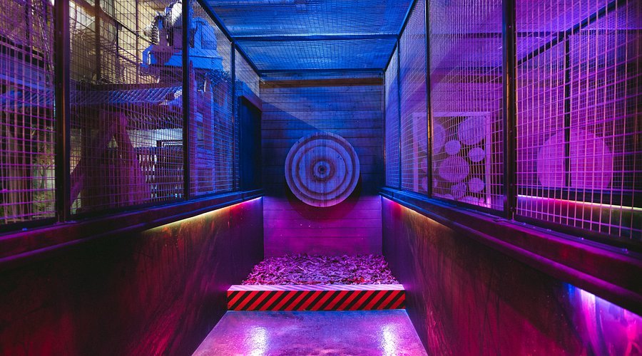An indoor axe-throwing lane with colorful purple and blue lighting, a wooden target on the far wall, and safety cages on either side.