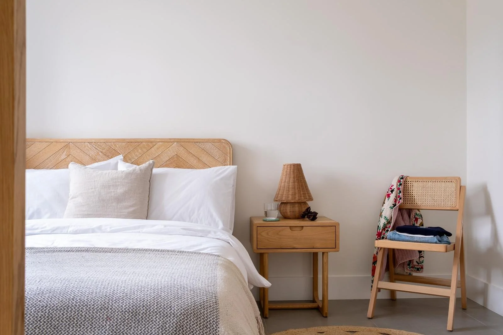 A neatly made bed with white pillows and a beige blanket in a minimalistic bedroom with light wood furniture, including a nightstand with a wicker lamp, a glass of water, and some clothes, and a wooden chair with folded clothes and a colorful towel.