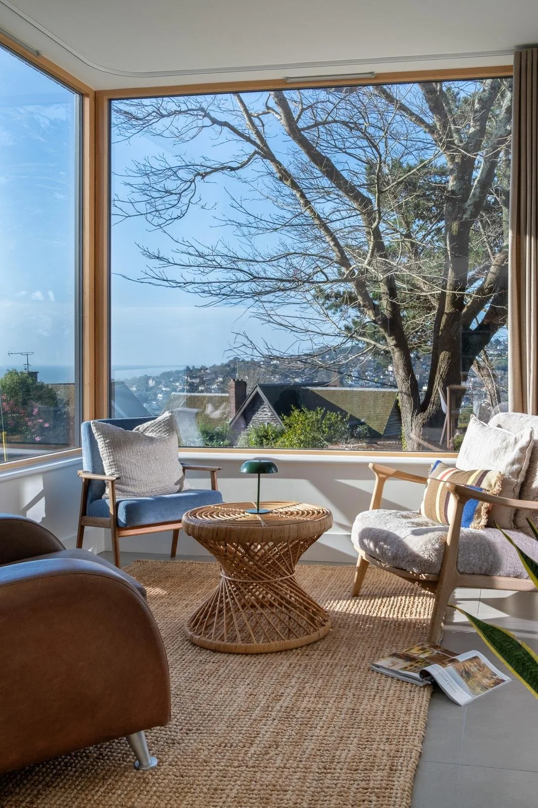 A cozy living room with a large window showcasing a view of leafless trees outside. The room contains a beige armchair, a wooden chair with light cushions, a woven rattan side table, and an open magazine on the floor. Sunlight illuminates the room, c
