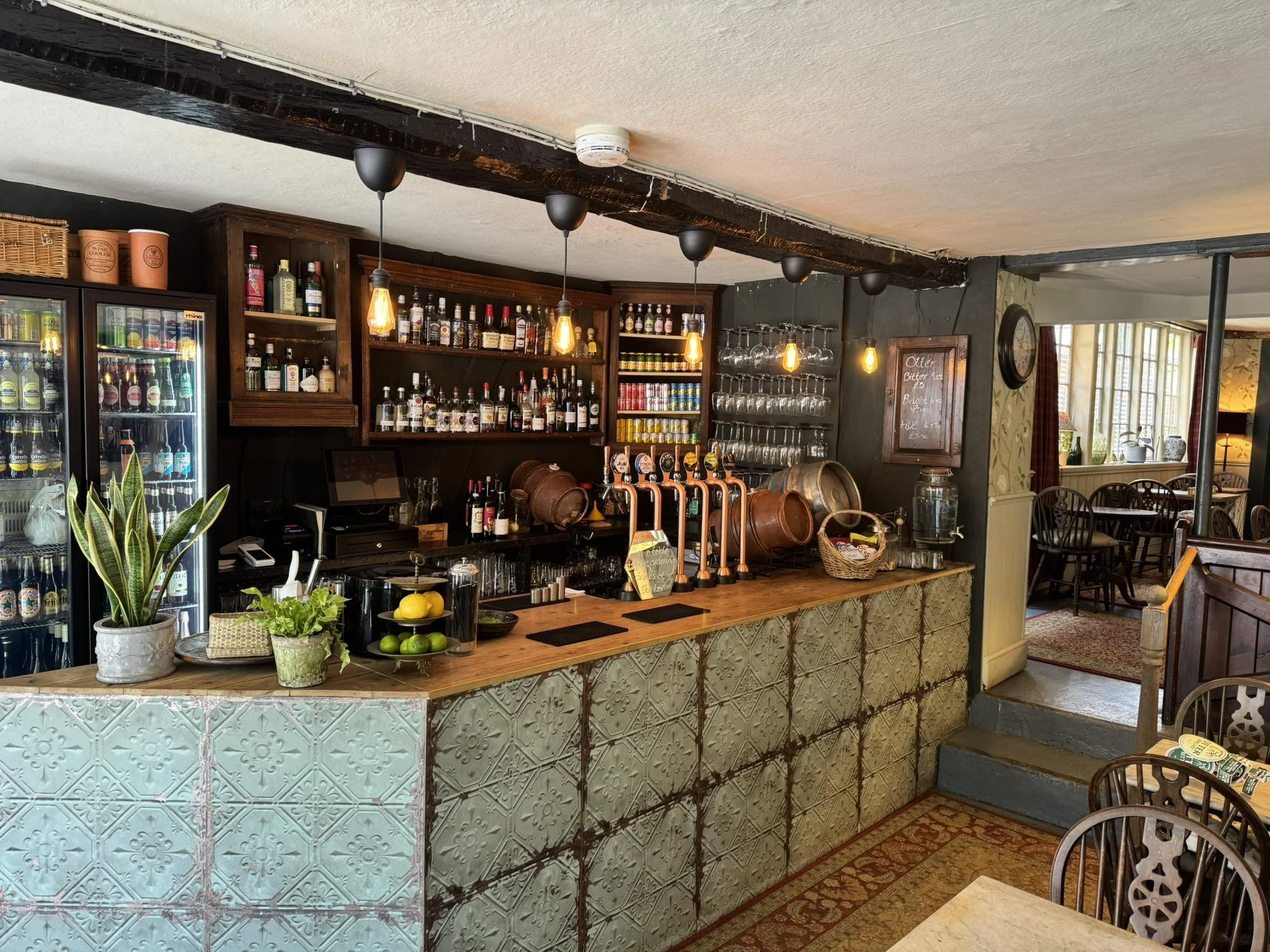 The image shows a cozy bar counter inside a restaurant or pub with shelves of bottles, glasses, and plants, with a doorway leading to a dining area in the background.