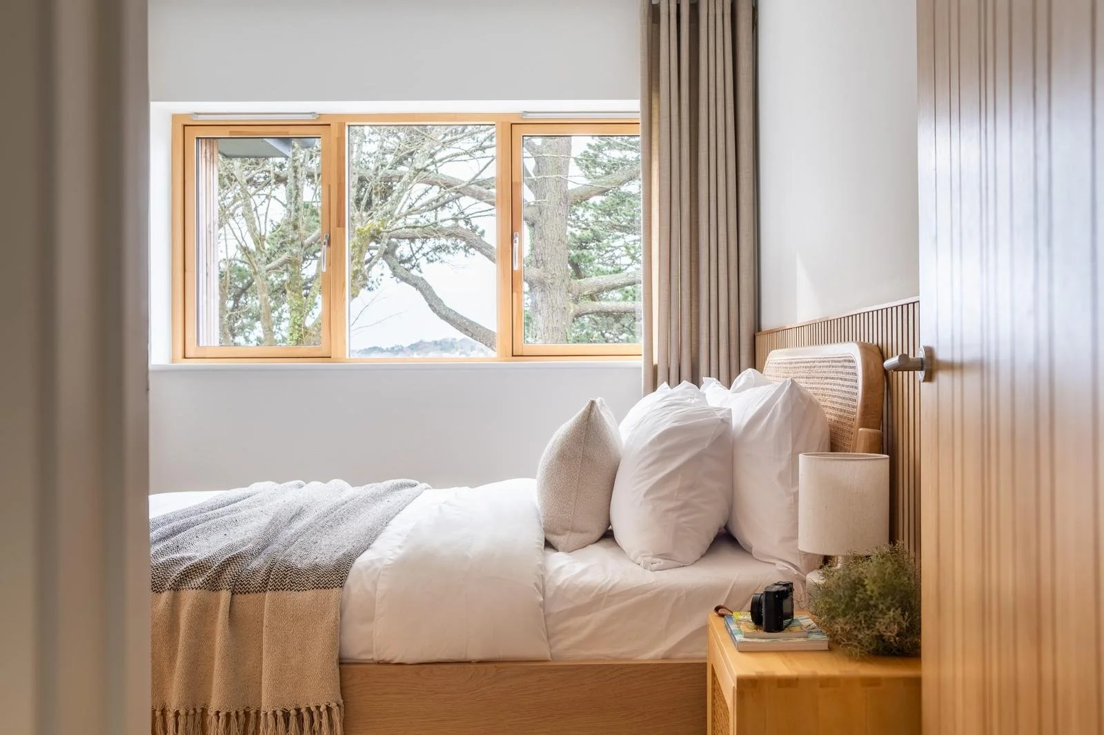 A cozy bedroom with a large wooden bed frame, white bedding, and multiple pillows. A nightstand with a small lamp, a camera, and a plant is next to the bed. Large windows with wooden frames show trees outside, allowing natural light into the room.